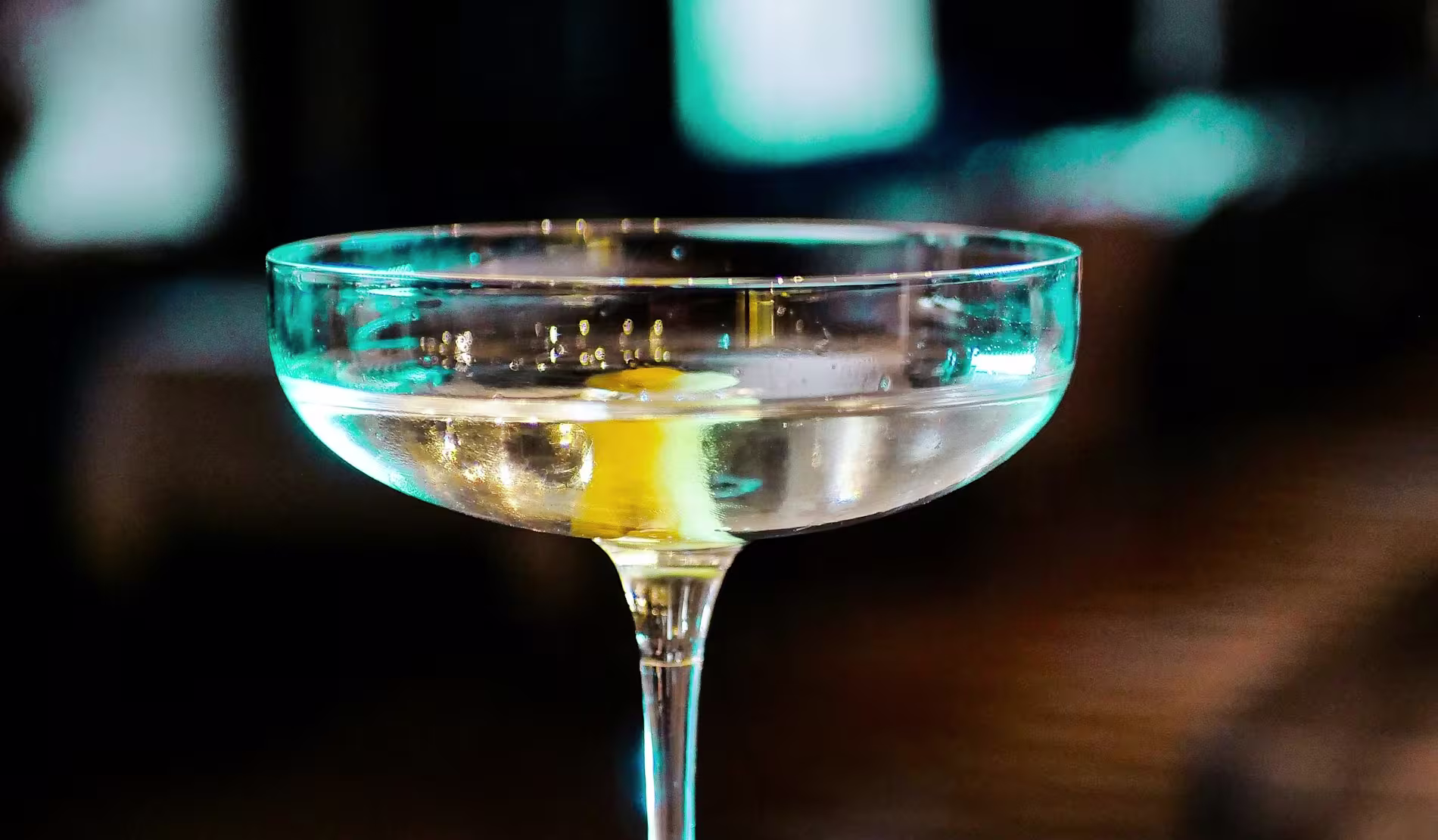 A close-up of a coupe glass filled with a clear cocktail garnished with a twist of lemon, with a blurred background.