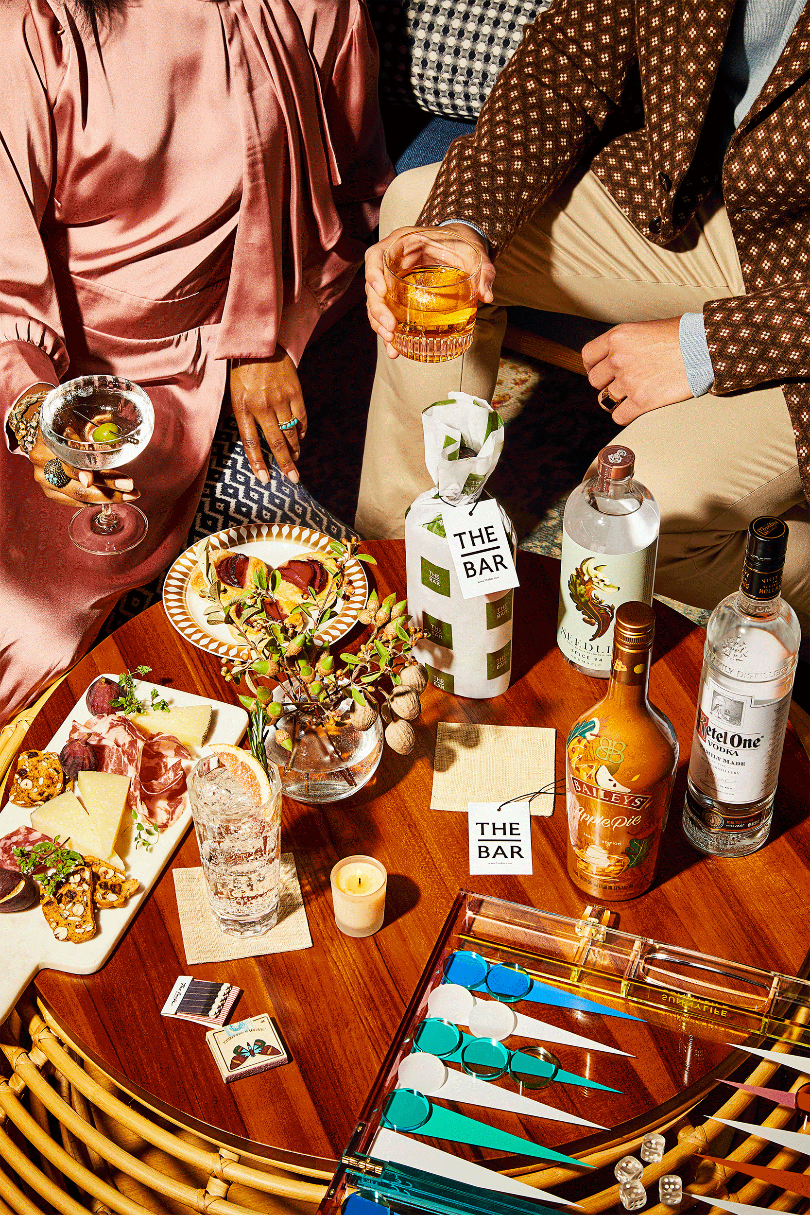A table with assorted cheeses, meats, cocktails, bottled spirits, and drinking glasses; two people are seated, each holding a drink.