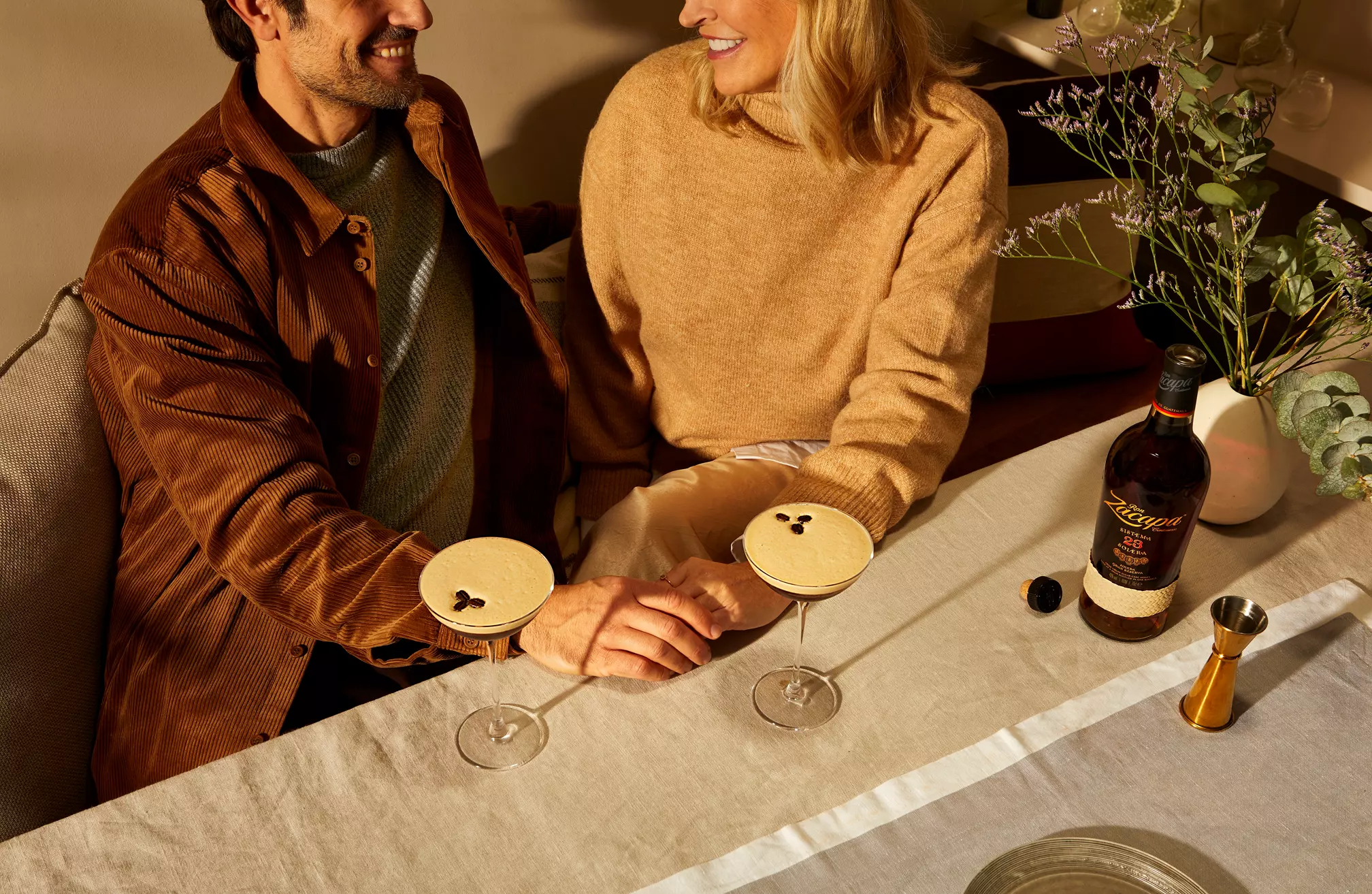 Man and woman sit at a table with two espresso martinis, holding hands and smiling. A bottle of rum, jigger, and flowers are also on the table.