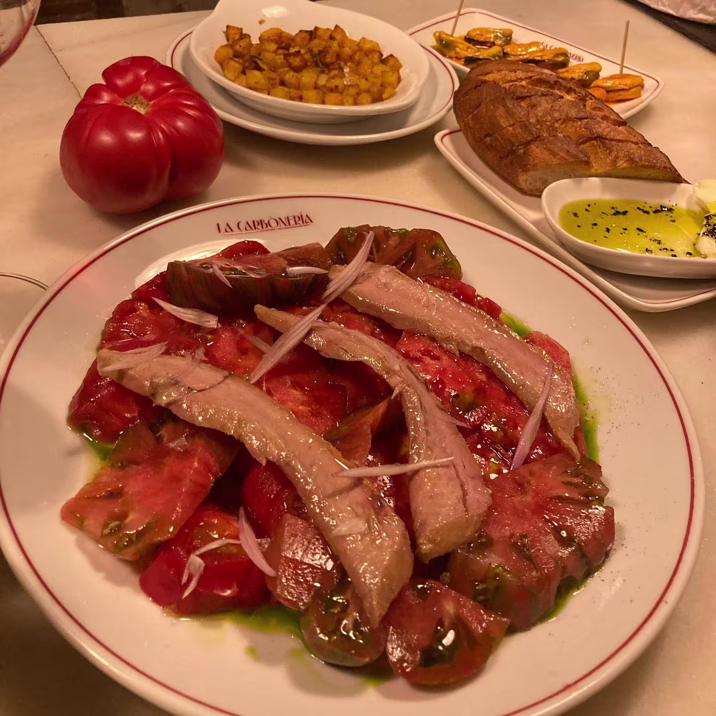 La foto muestra platos de la carta del restaurante La Carbonería, los platos expuestos son unos tomates con ventresca, unas migas y unos mejillones.