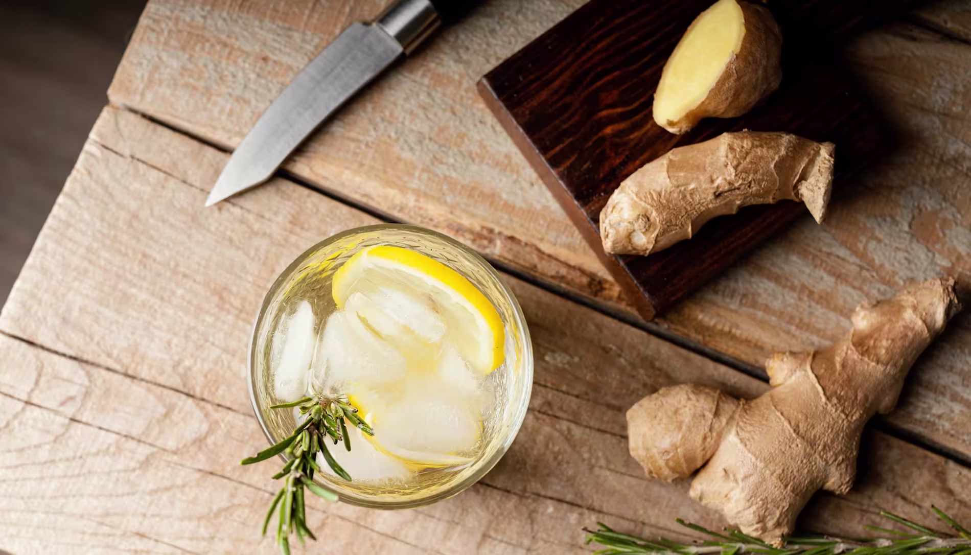 A glass of iced drink with lemon and rosemary sits on a wooden table next to fresh ginger, a cutting board, and a knife.