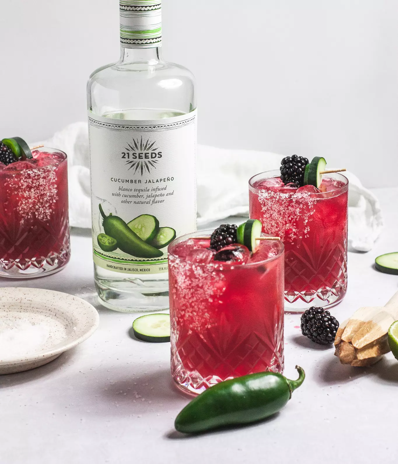 A bottle of 21 Seeds Cucumber Jalapeño tequila behind two glasses of a red cocktail garnished with blackberries, cucumber slices, and jalapeño, with salt and jalapeño on the table.