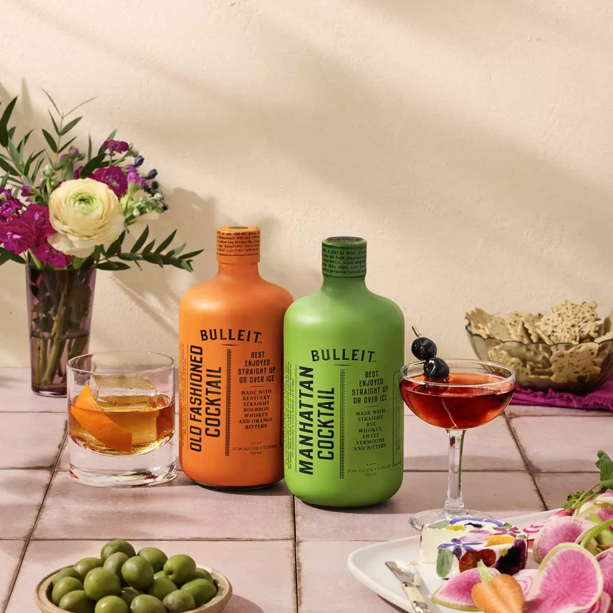 Two colorful Bulleit cocktail bottles, Old Fashioned (orange) and Manhattan (green), sit on a tiled table with garnished drinks, flowers, and snacks in a bright setting.