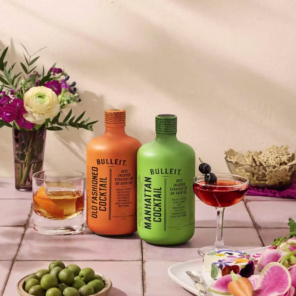 Two colorful Bulleit cocktail bottles, Old Fashioned (orange) and Manhattan (green), sit on a tiled table with garnished drinks, flowers, and snacks in a bright setting.