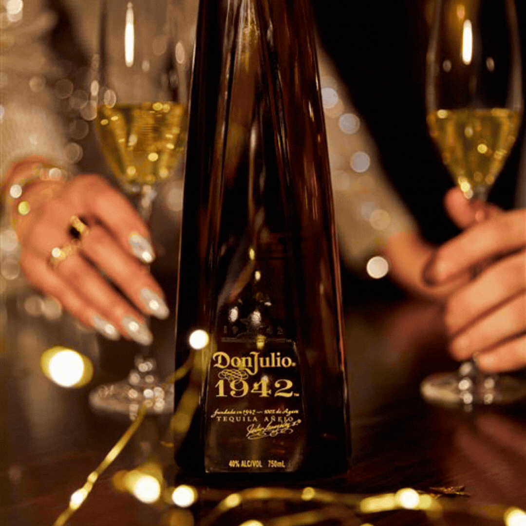 A bottle of Don Julio 1942 tequila is placed on a table between two people holding champagne glasses, with festive string lights in the foreground.