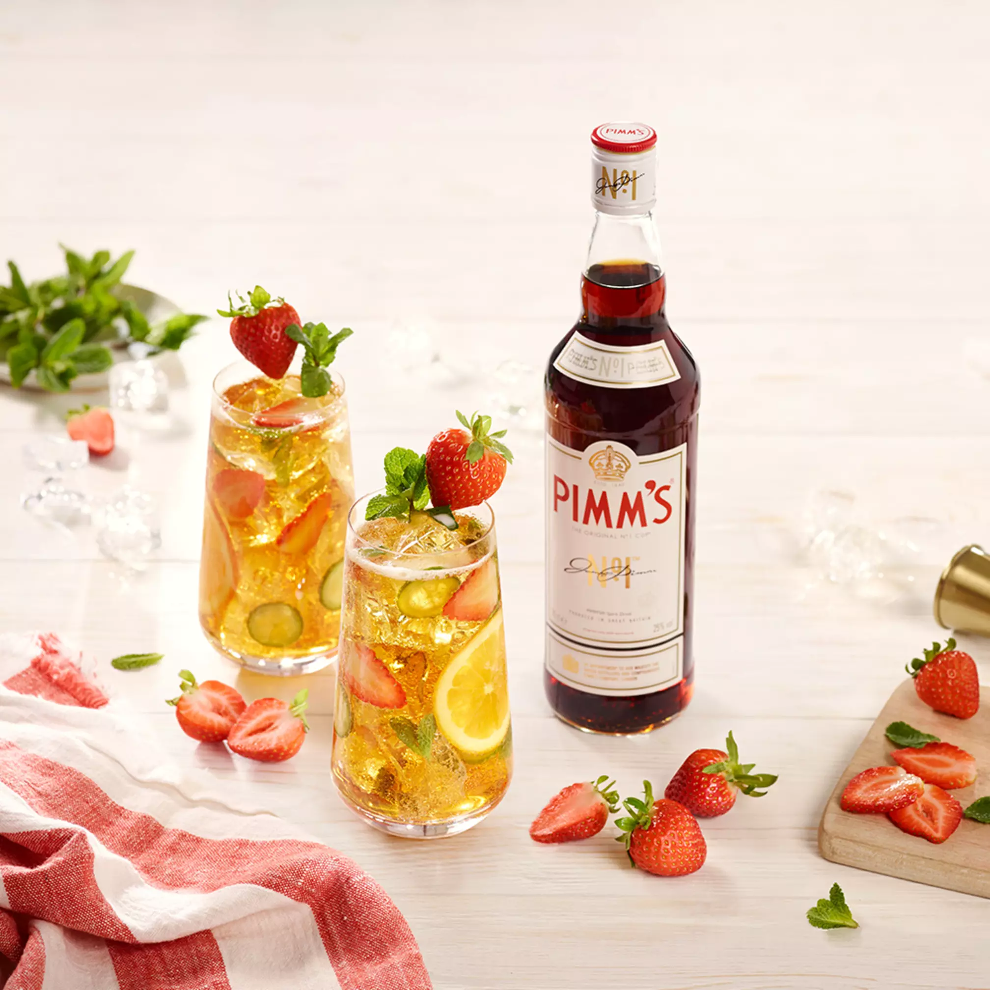 A bottle of Pimm’s No. 1 is shown next to two glasses of Pimm’s cocktail with ice, lemon, cucumber, strawberries, and mint, on a light surface with strawberries and a red-striped cloth.