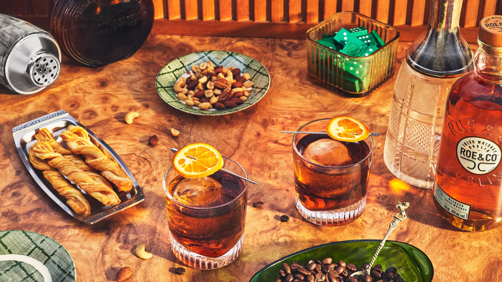 Two cocktails with orange garnishes, a bottle of Rye whiskey, mixed nuts, breadsticks, a cocktail shaker, coffee beans, and a dish of dice are arranged on a wooden table.