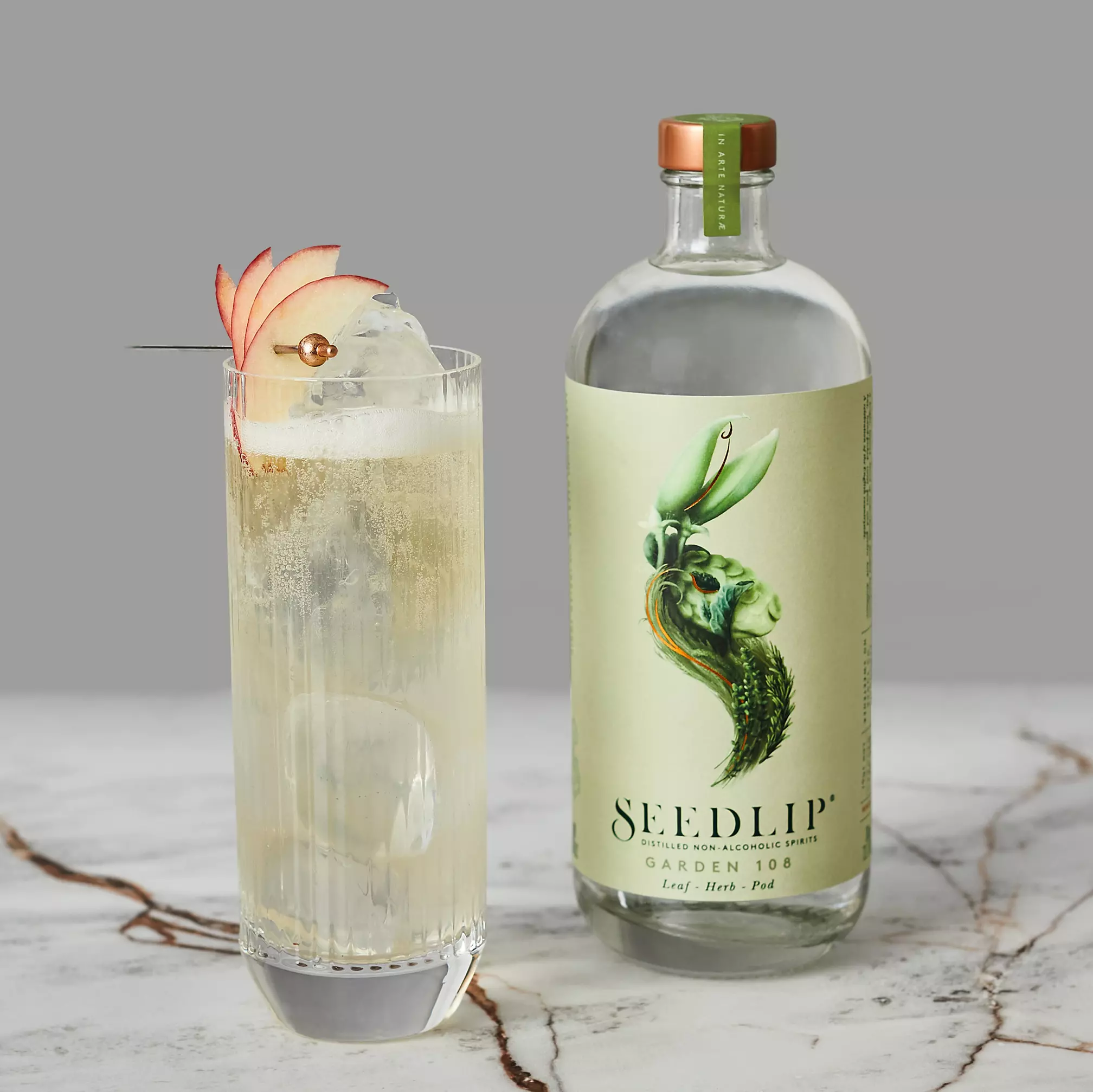 A tall glass with a light-colored drink, ice, and apple slices on a pick sits next to a bottle of Seedlip Garden 108 on a marble surface with a gray background.