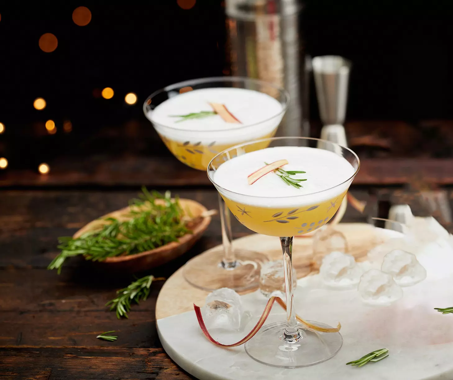 Two cocktails with frothy tops, garnished with rosemary and citrus peel, sit on a marble board with ice cubes and herb sprigs on a rustic wooden table.