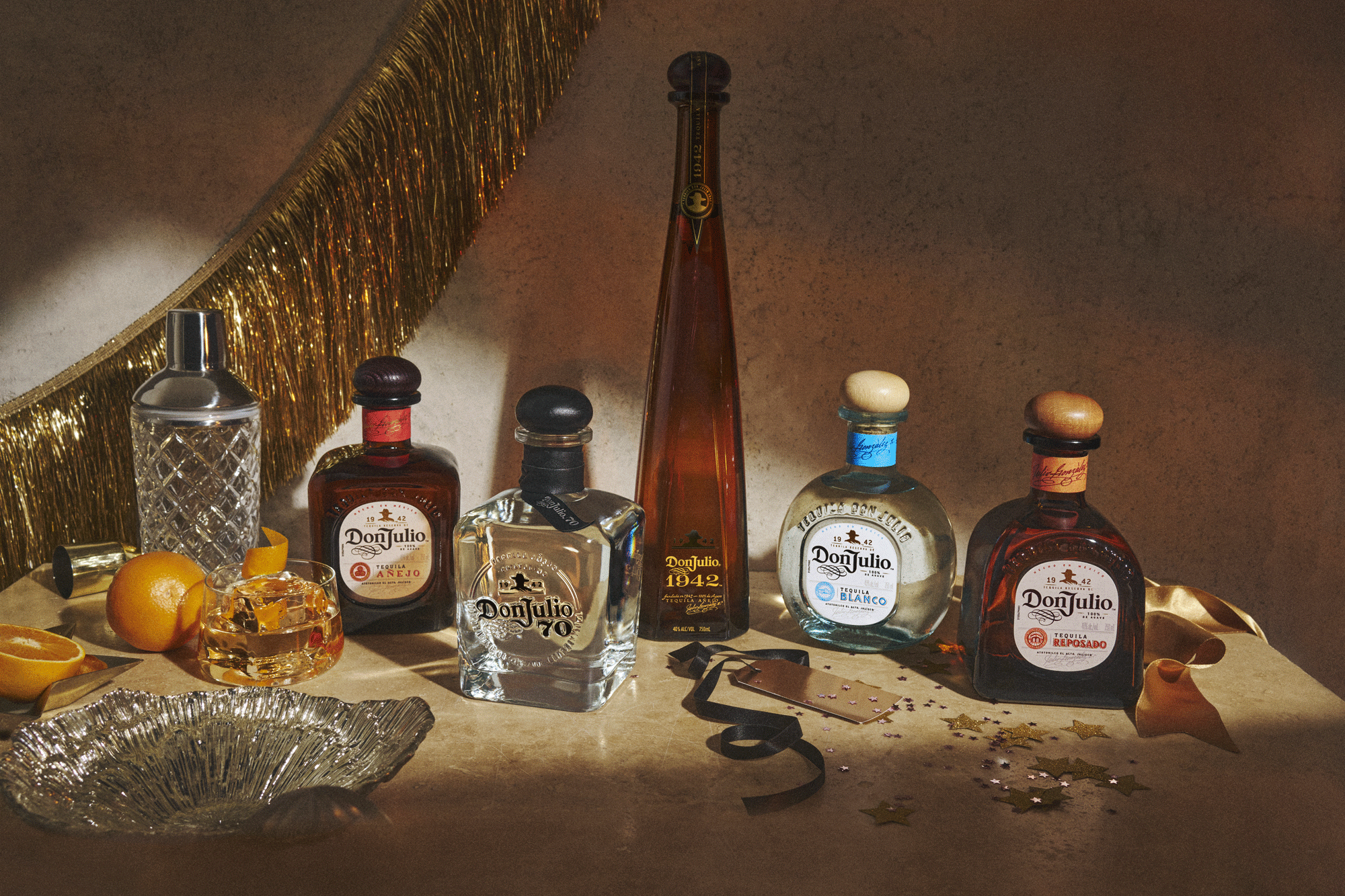 A selection of Don Julio tequila bottles and a Don Julio 1942 bottle are arranged on a table with a cocktail, shaker, oranges, glassware, and gold party decorations.