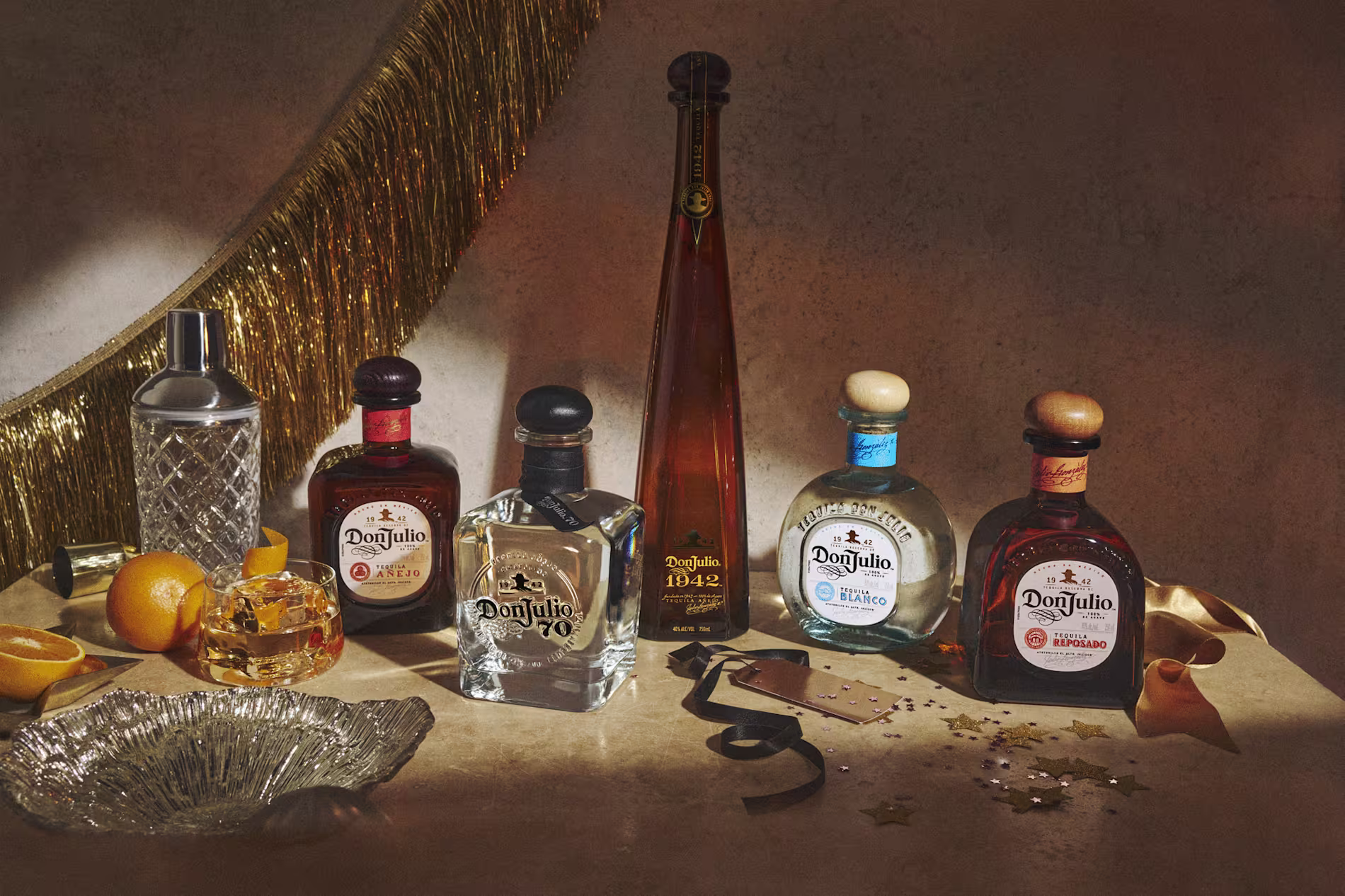 A selection of Don Julio tequila bottles and a Don Julio 1942 bottle are arranged on a table with a cocktail, shaker, oranges, glassware, and gold party decorations.