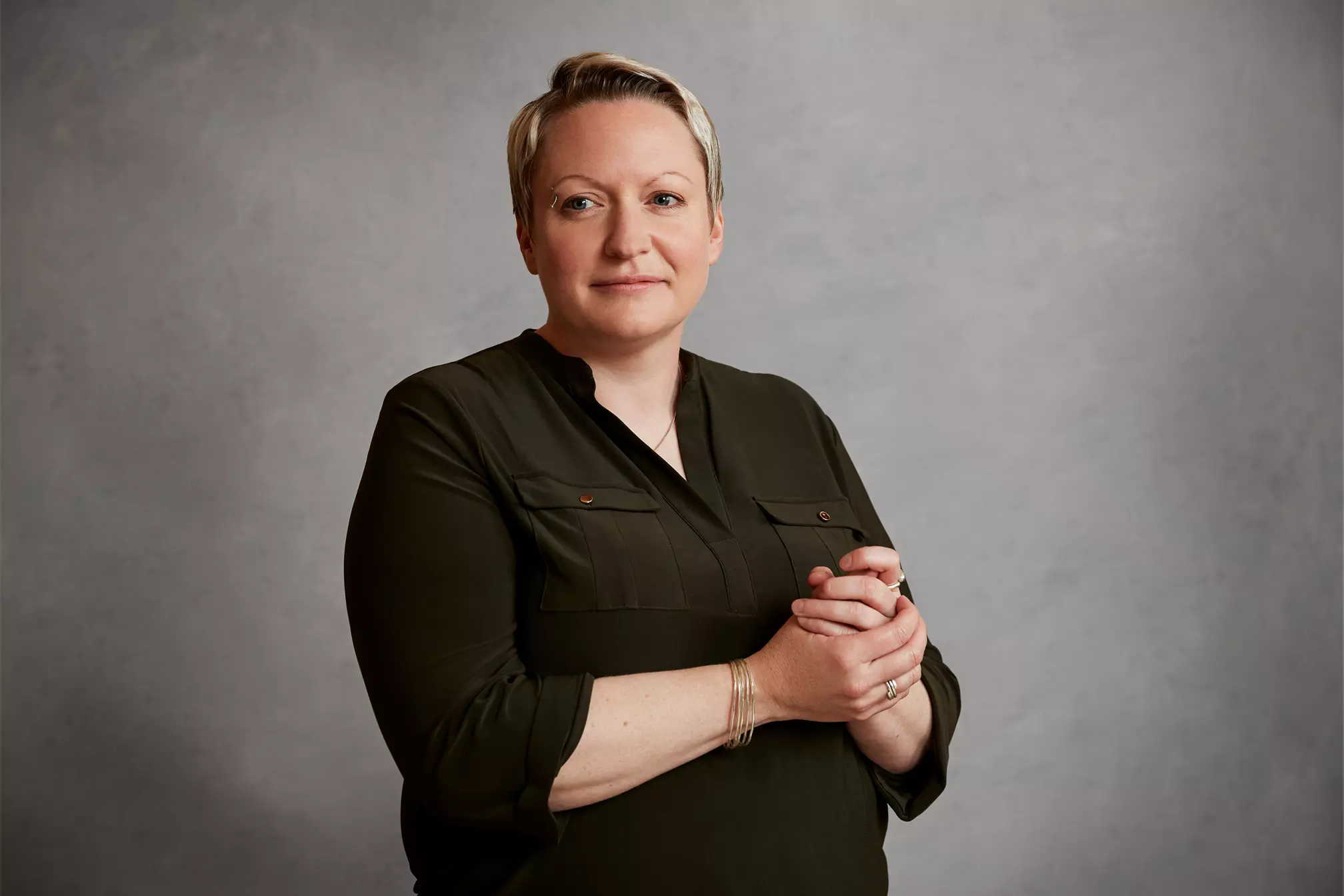 A person with short blond hair wearing a dark long-sleeved top stands against a plain gray background, looking slightly to the side with hands clasped.