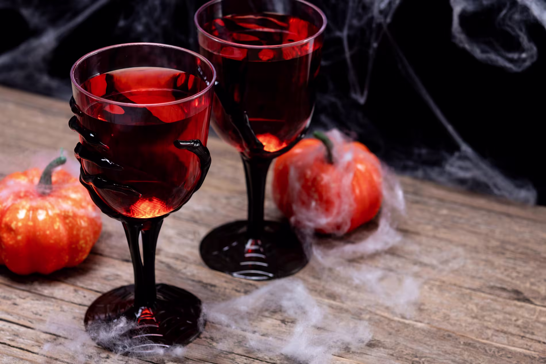 Two Halloween-themed wine glasses with black hand-shaped stems filled with red liquid, surrounded by artificial pumpkins and cobwebs on a wooden surface.