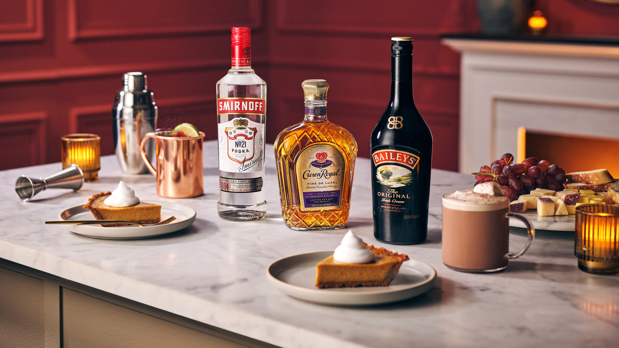 Bottles of Smirnoff vodka, Crown Royal whisky, and Baileys Irish Cream on a marble counter with cocktails, pumpkin pie slices, and candles.