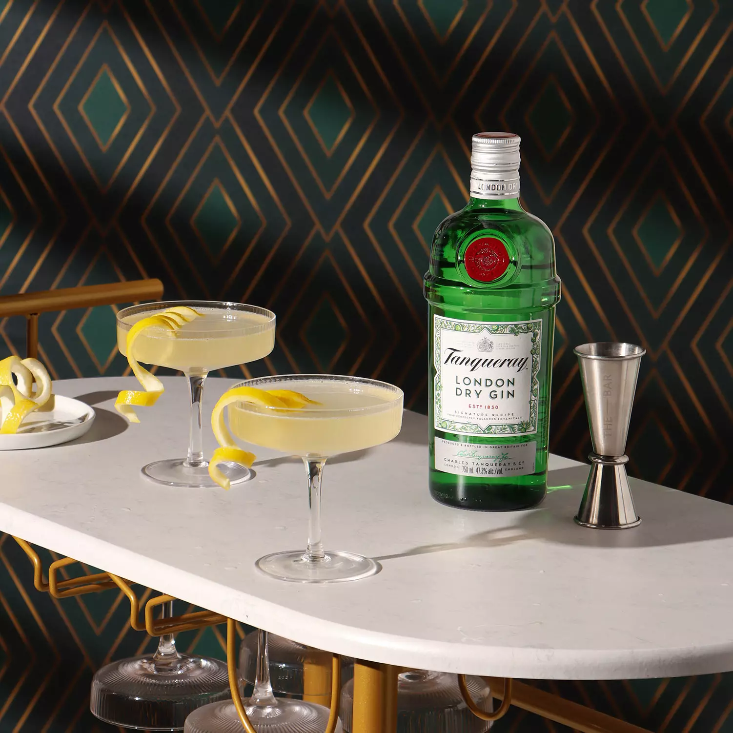 A bottle of Tanqueray London Dry Gin stands on a marble surface next to two cocktails with lemon twists and a metal jigger, set against a geometric patterned background.