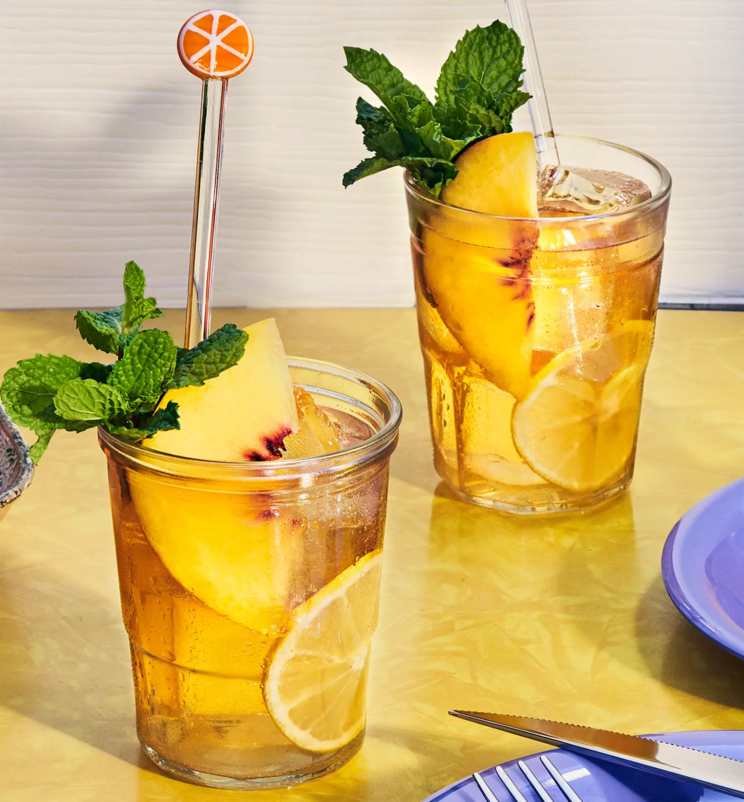 Two glasses of iced tea garnished with lemon slices, peach wedges, and mint sprigs sit on a yellow table next to a blue plate and silver cutlery.