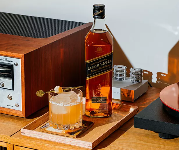A bottle of Johnnie Walker Black Label whisky next to a glass of cocktail with ice and garnish, placed on a wooden tray beside audio equipment.