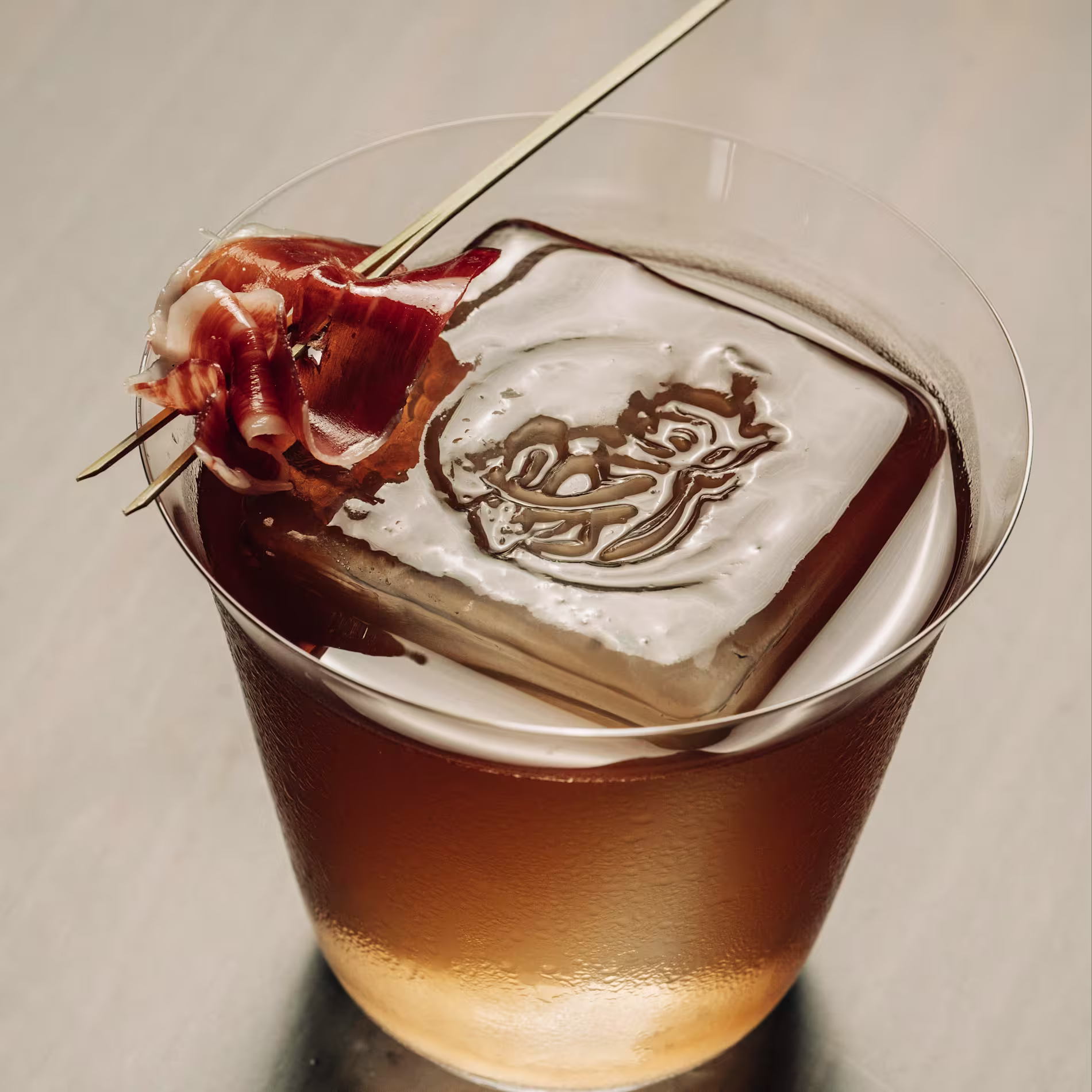Cóctel ámbar con gran cubo de hielo grabado y virutas de jamón ibérico como garnish sobre un pincho dorado.