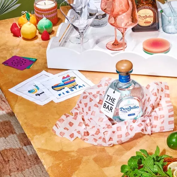 A bottle of Don Julio tequila sits on a patterned tablecloth beside cocktail napkins, glassware, limes, and a tray of liquor bottles on a colorful table setup.