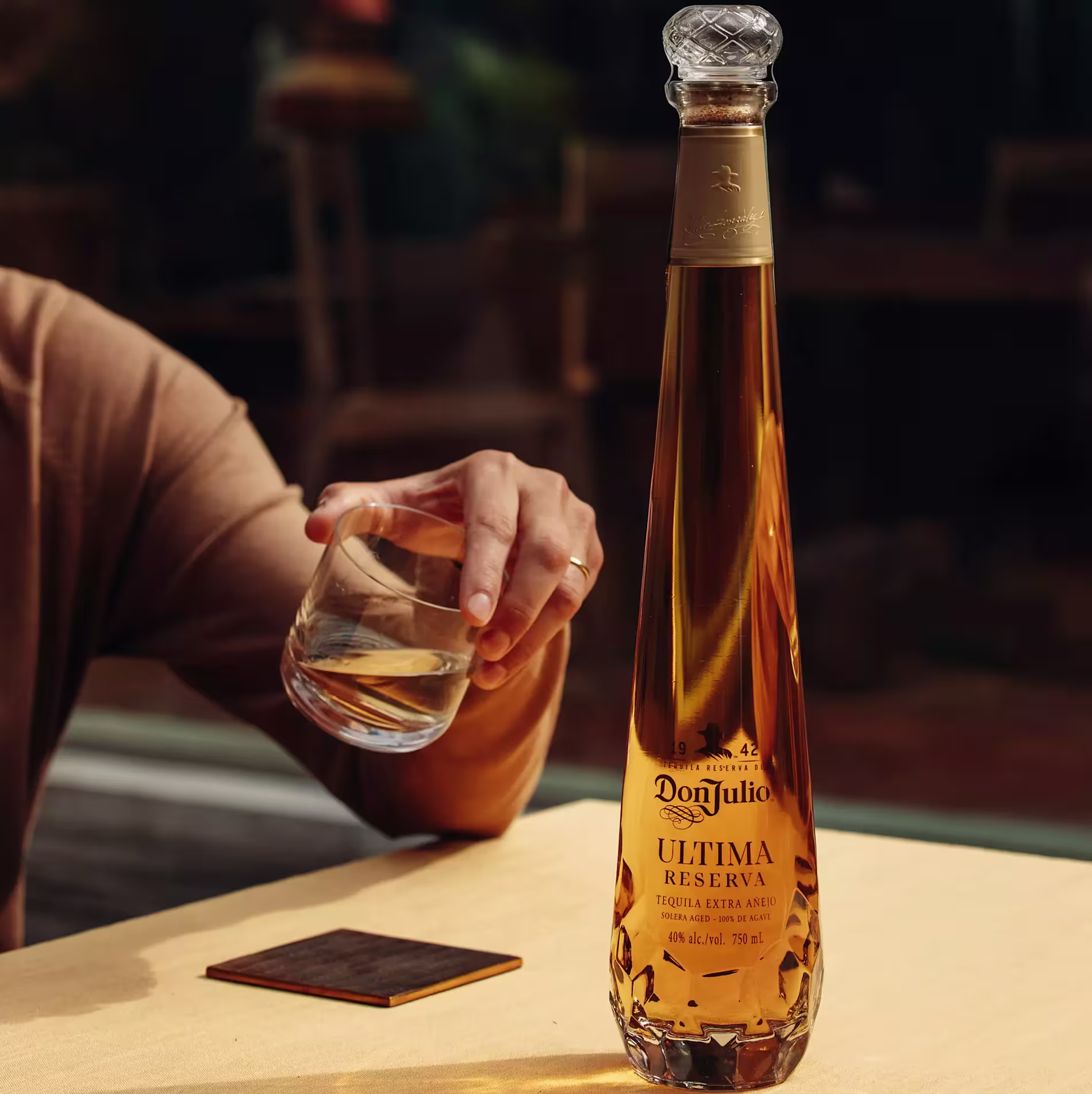 A person holds a glass of clear liquid next to a bottle of Don Julio Ultima Reserva tequila on a table with a coaster.