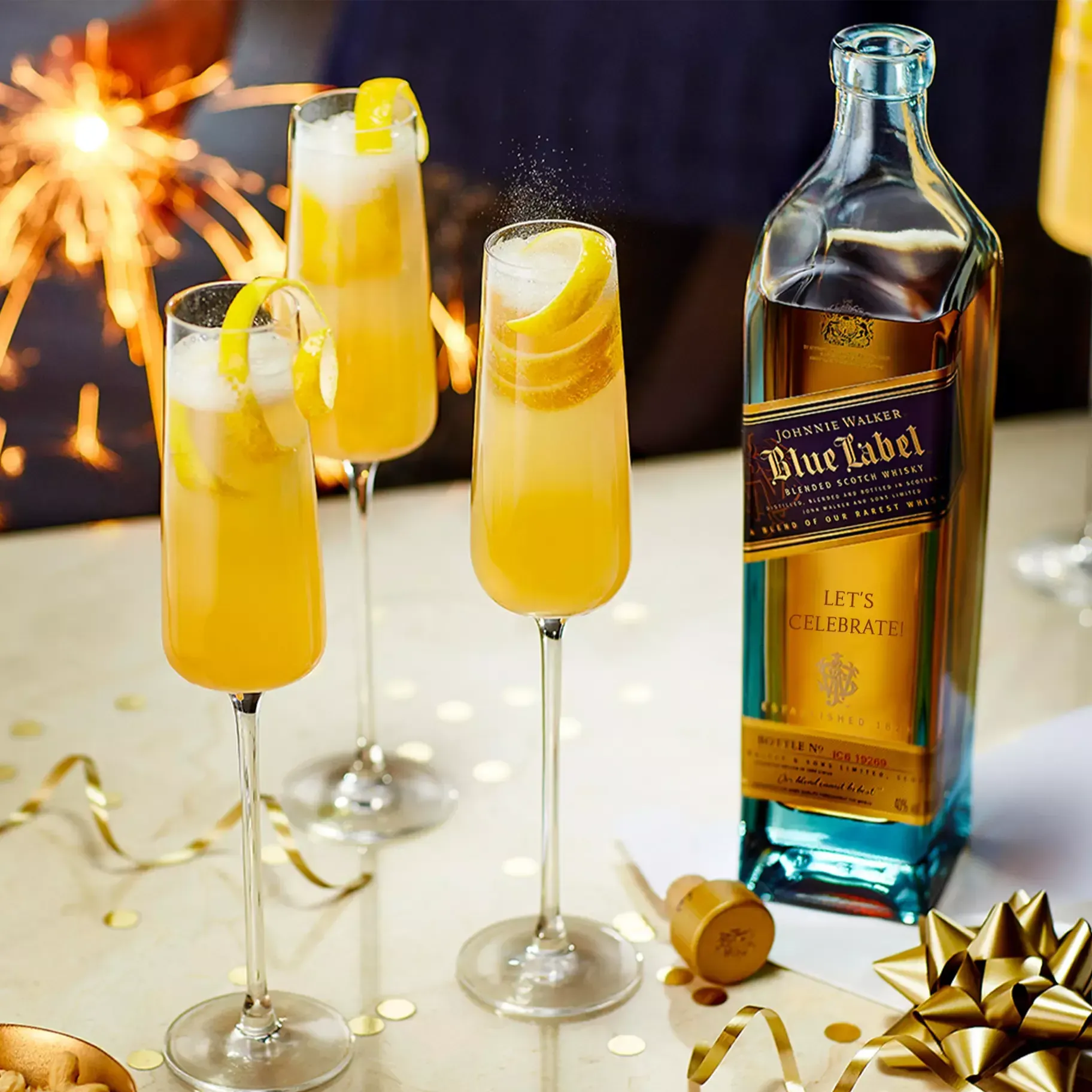A bottle of Johnnie Walker Blue Label with three champagne flutes of orange cocktails garnished with lemon, surrounded by party decorations and a sparkler in the background.