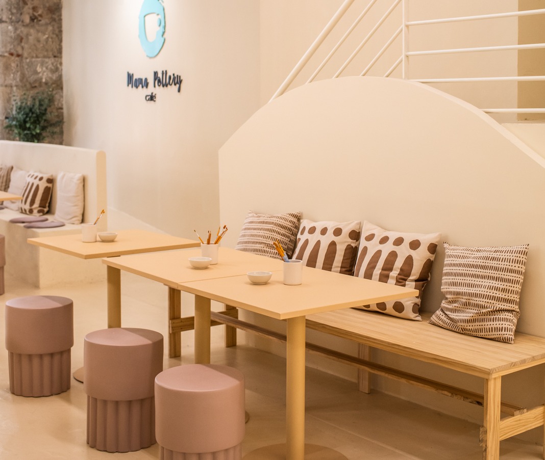 Zona de mesas en Mama Pottery Café con bancos de madera, taburetes redondos color malva, cojines estampados y materiales listos para pintar cerámica.