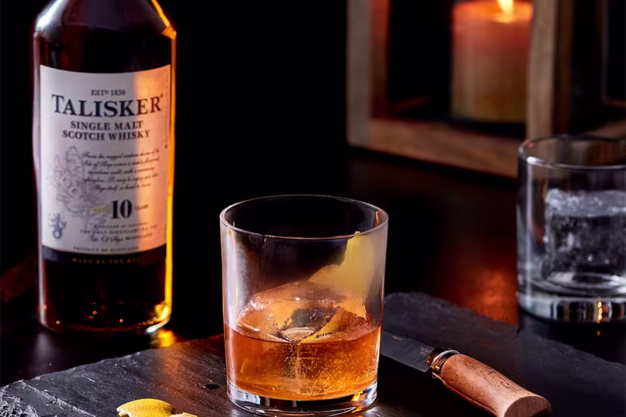 A glass of whisky with a lemon twist sits on a dark slate next to a bottle of Talisker 10 Year Single Malt Scotch, a knife, and a candle in the background.
