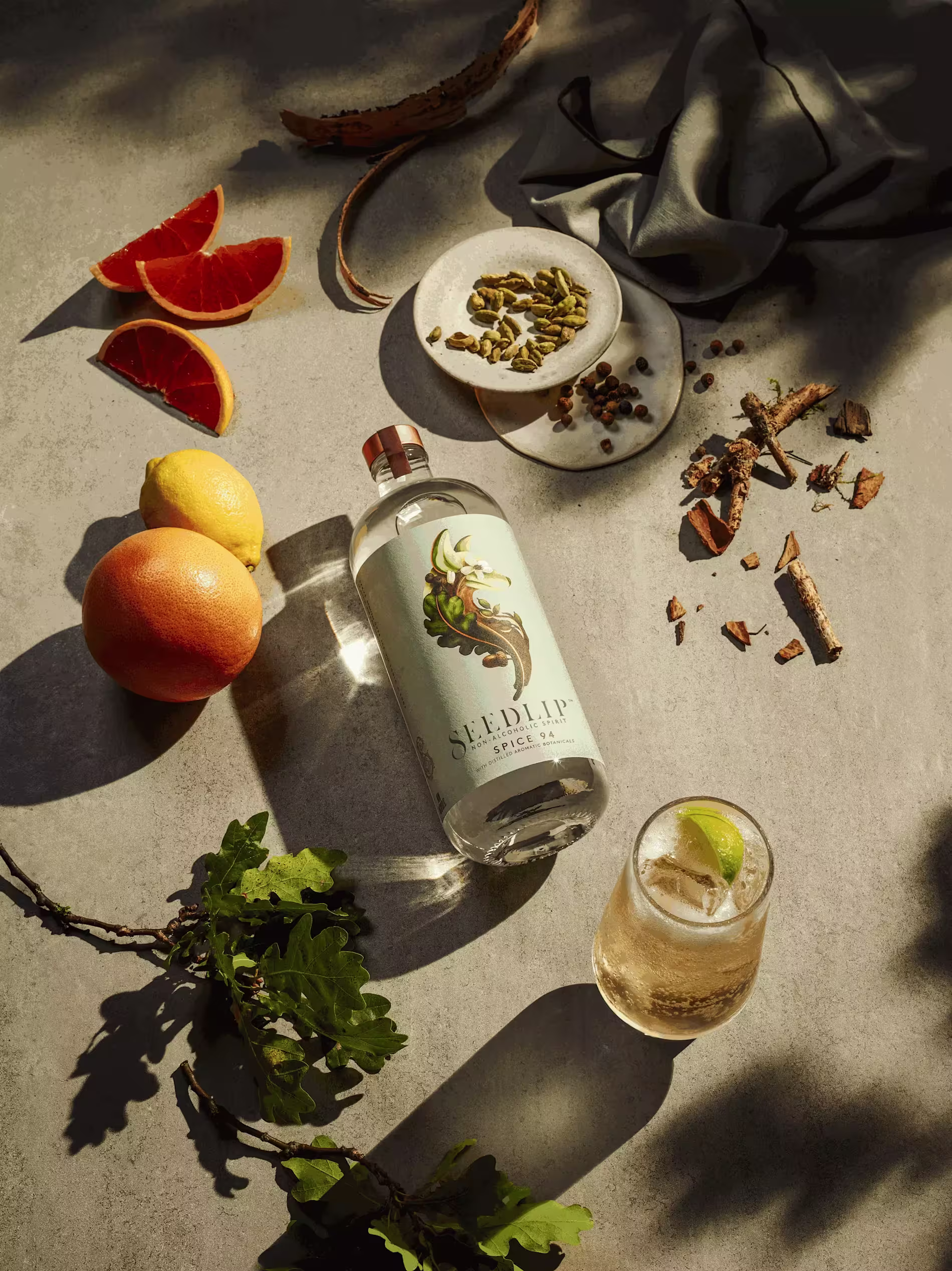 A bottle of Seedlip Spice 94, a glass with a lime wedge, grapefruit halves, spices, and greenery arranged on a textured surface.