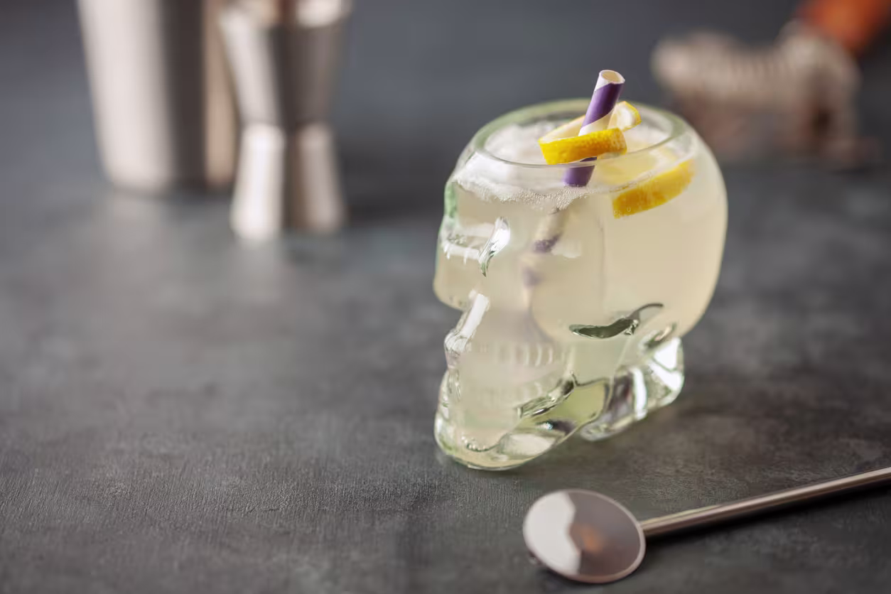 A cocktail with lemon slices and a straw is served in a glass shaped like a skull, placed on a dark surface next to a metal spoon and bar tools.