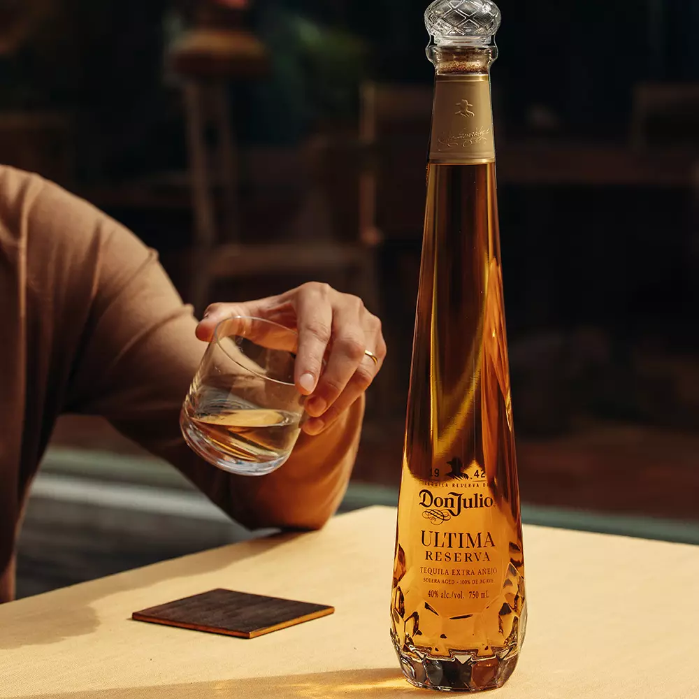 A person holding a glass of clear liquid next to a tall bottle of Don Julio Ultima Reserva tequila on a table.