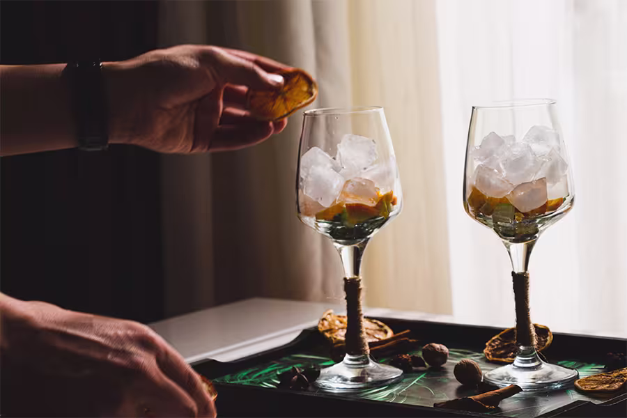 A person places a dried citrus slice into a wine glass filled with ice; another similar glass is beside it on a tray with assorted dried fruits and spices.