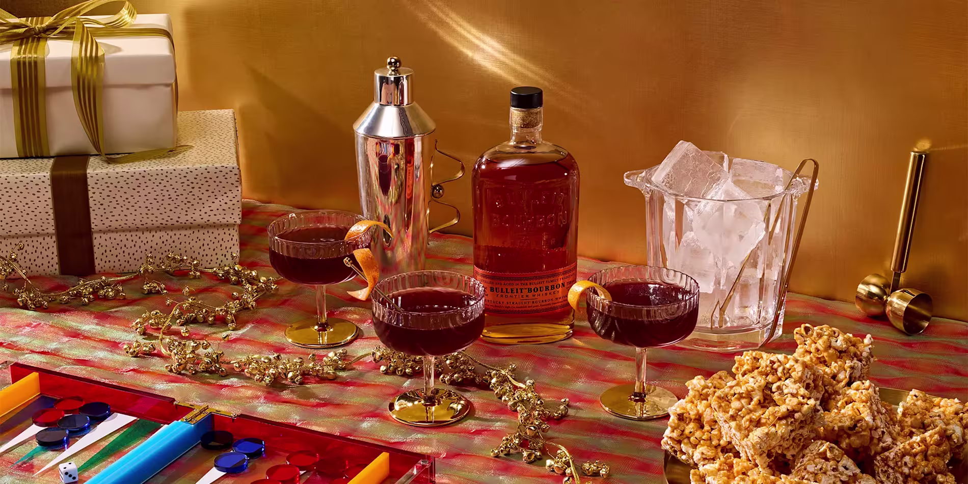 A festive table with three cocktails, a bottle of Bulleit Bourbon, a cocktail shaker, an ice bucket, popcorn, gift boxes, and a backgammon game on a striped tablecloth.