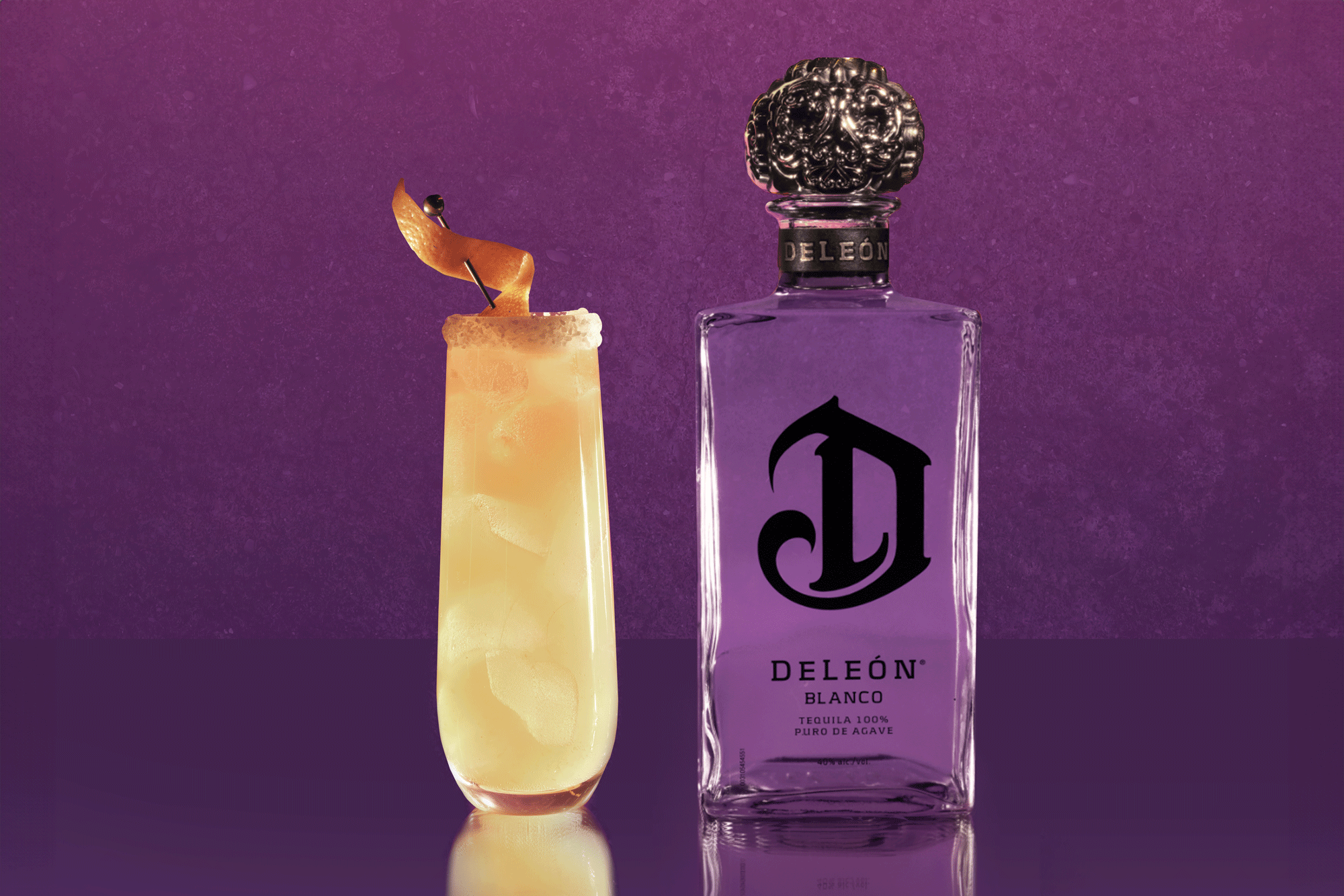 A tall cocktail garnished with an orange twist sits next to a bottle of DeLeón Blanco tequila against a purple background.