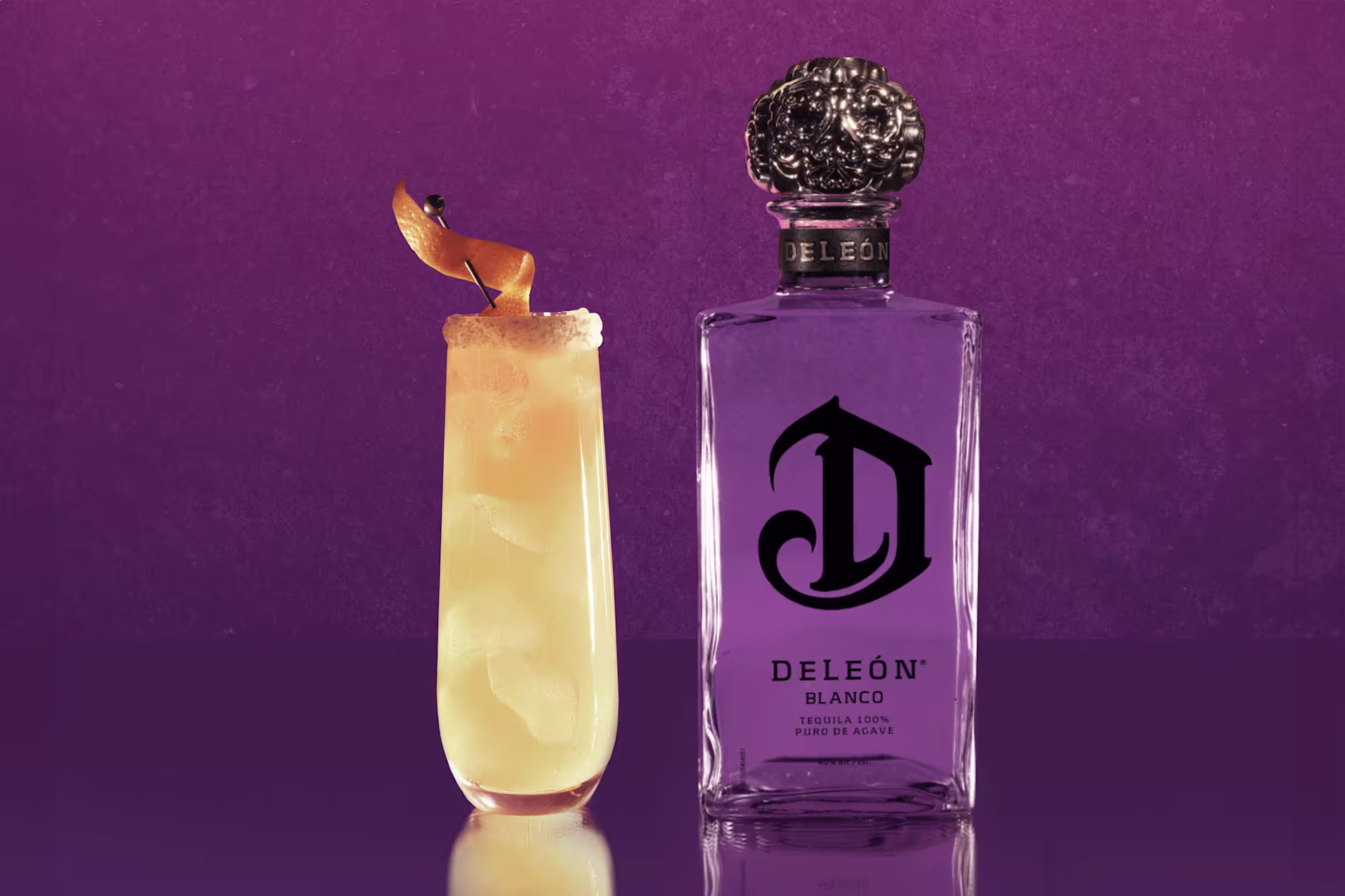 A tall cocktail garnished with an orange twist sits next to a bottle of DeLeón Blanco tequila against a purple background.