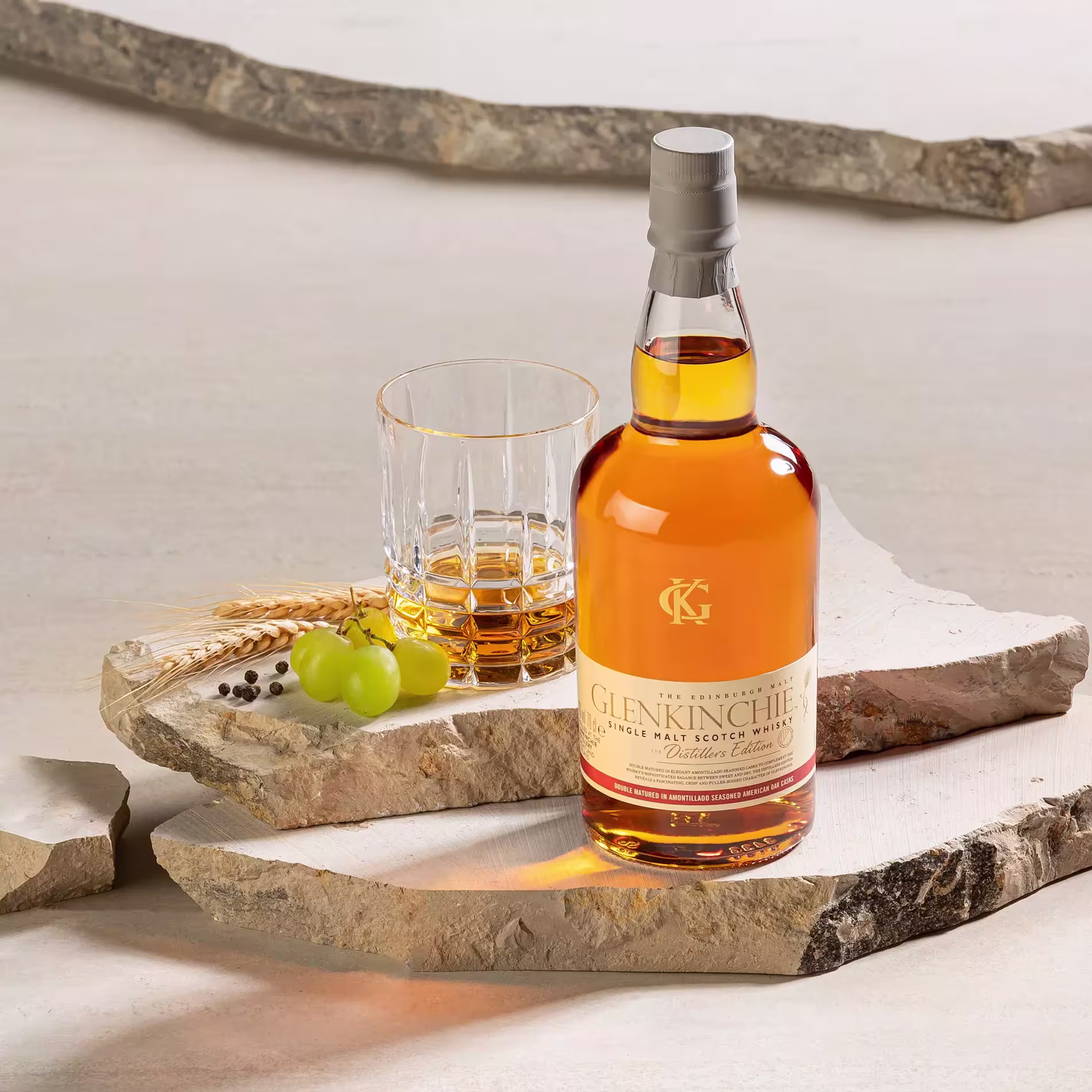 A bottle of Glenkinchie single malt Scotch whisky next to a filled glass, grapes, barley, and peppercorns, all arranged on stone slabs.