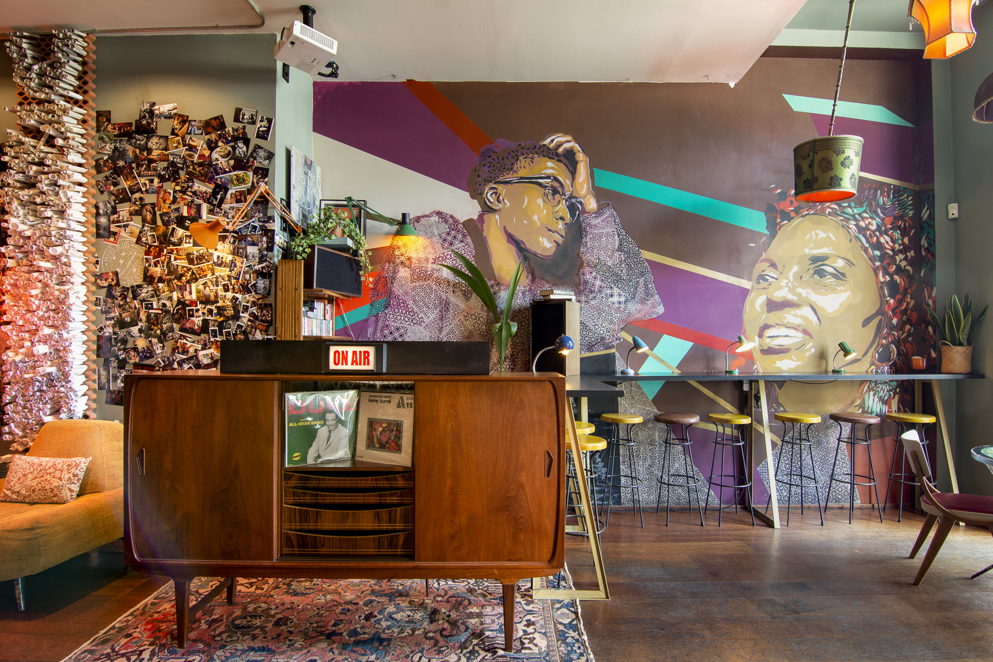 La foto muestra el espacio interior de Guzzon. Contiene discos y un tocadiscos. Detrás, hay una zona de bar con taburetes y luces colgantes. Las paredes están decoradas con patrones geométricos coloridos y un collage de numerosas fotos pequeñas.