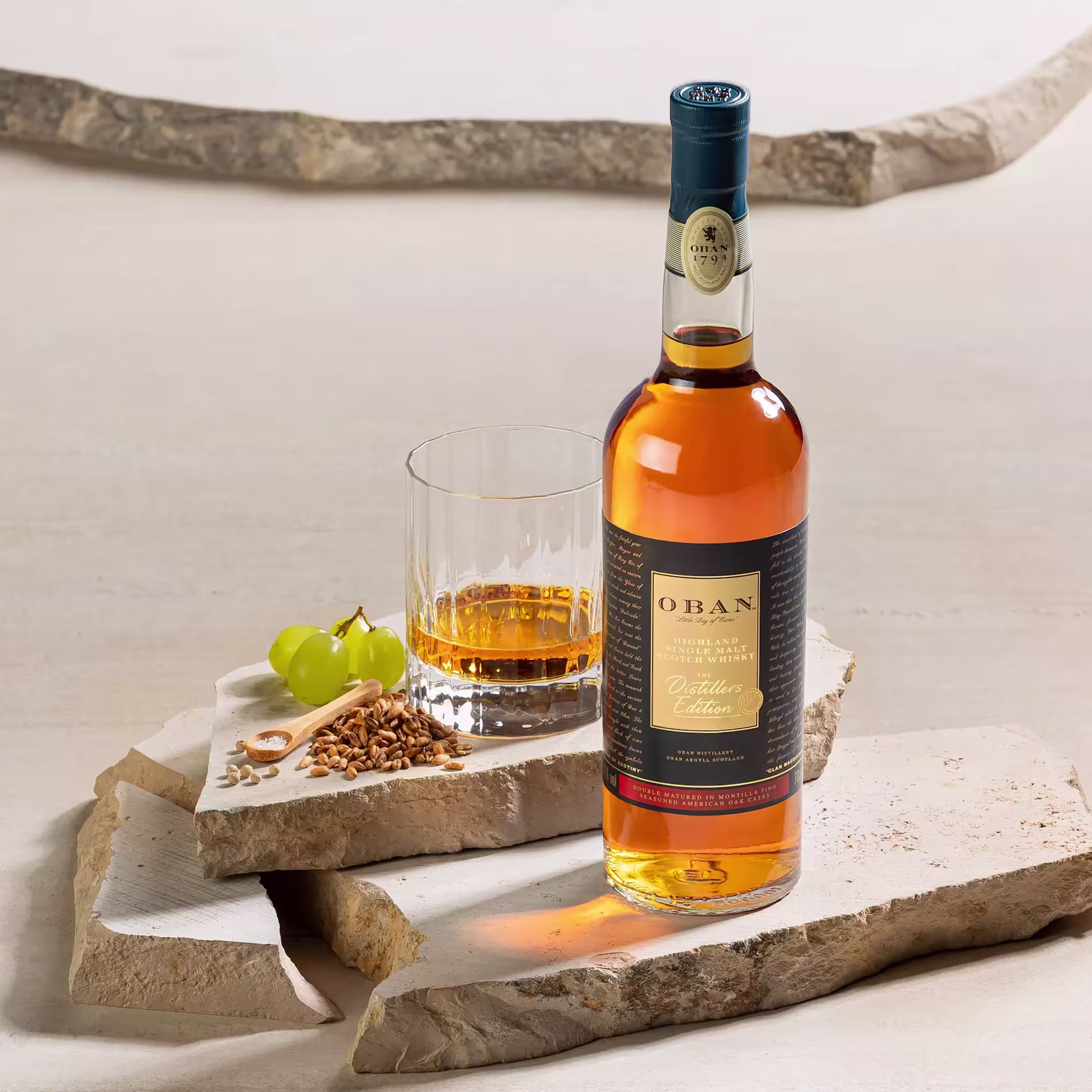 A bottle of Oban Distillers Edition whisky stands beside a glass of whisky, green grapes, and grains on stone slabs.