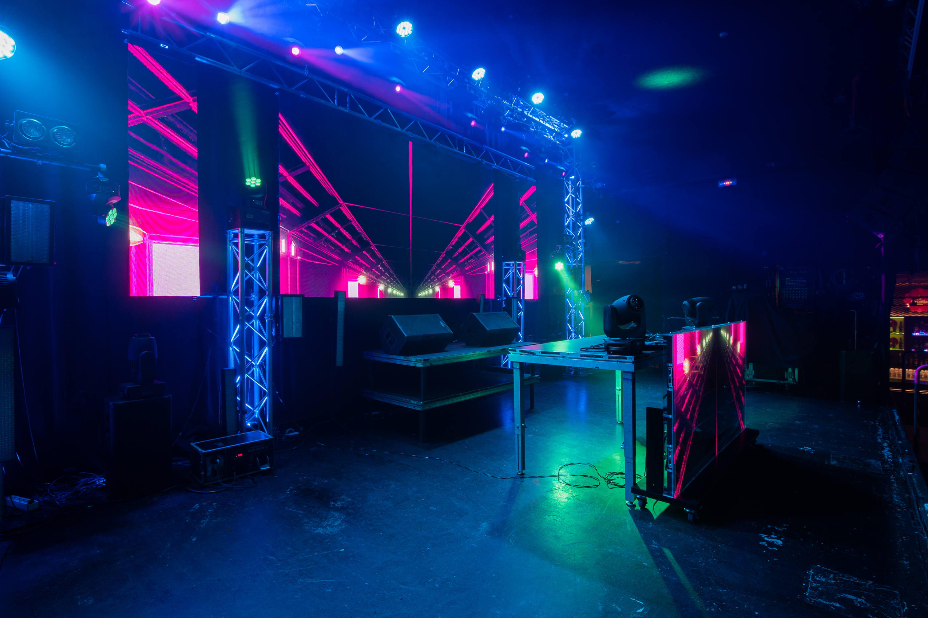 La imagen muestra el escenario de una sala de conciertos con luces de neón y pantallas LED en tonos vibrantes de azul, rosa y verde. El espacio está diseñado para crear una experiencia visual inmersiva y energética para el público.