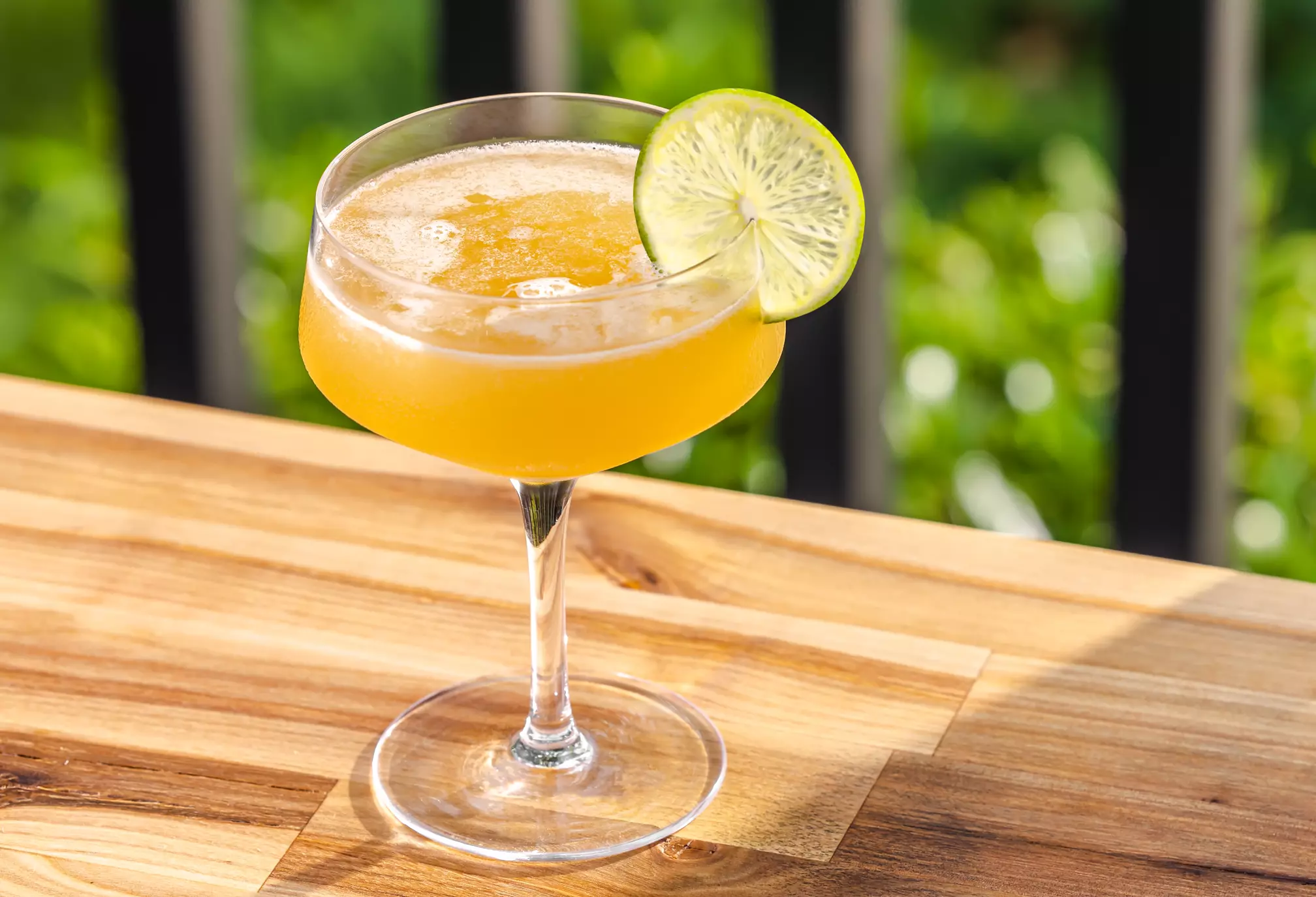 A yellow cocktail served in a coupe glass with a lime wheel garnish, placed on a wooden table outdoors.