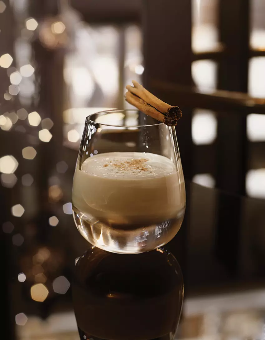 A glass of creamy cocktail topped with ground spice and garnished with two cinnamon sticks, set on a reflective surface with blurred lights in the background.