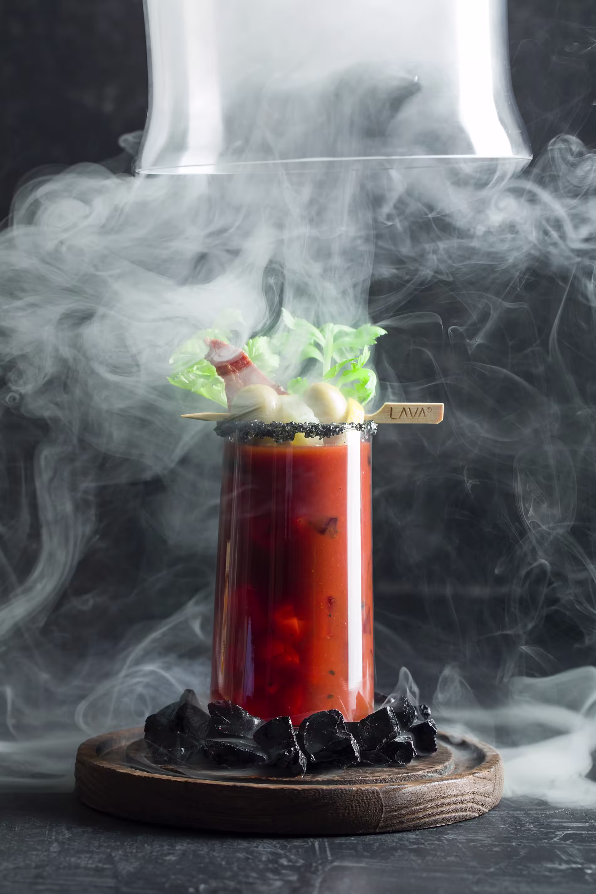 A tall glass of red cocktail with leafy garnishes and black salt rim sits on a wooden base surrounded by black stones and swirling smoke.
