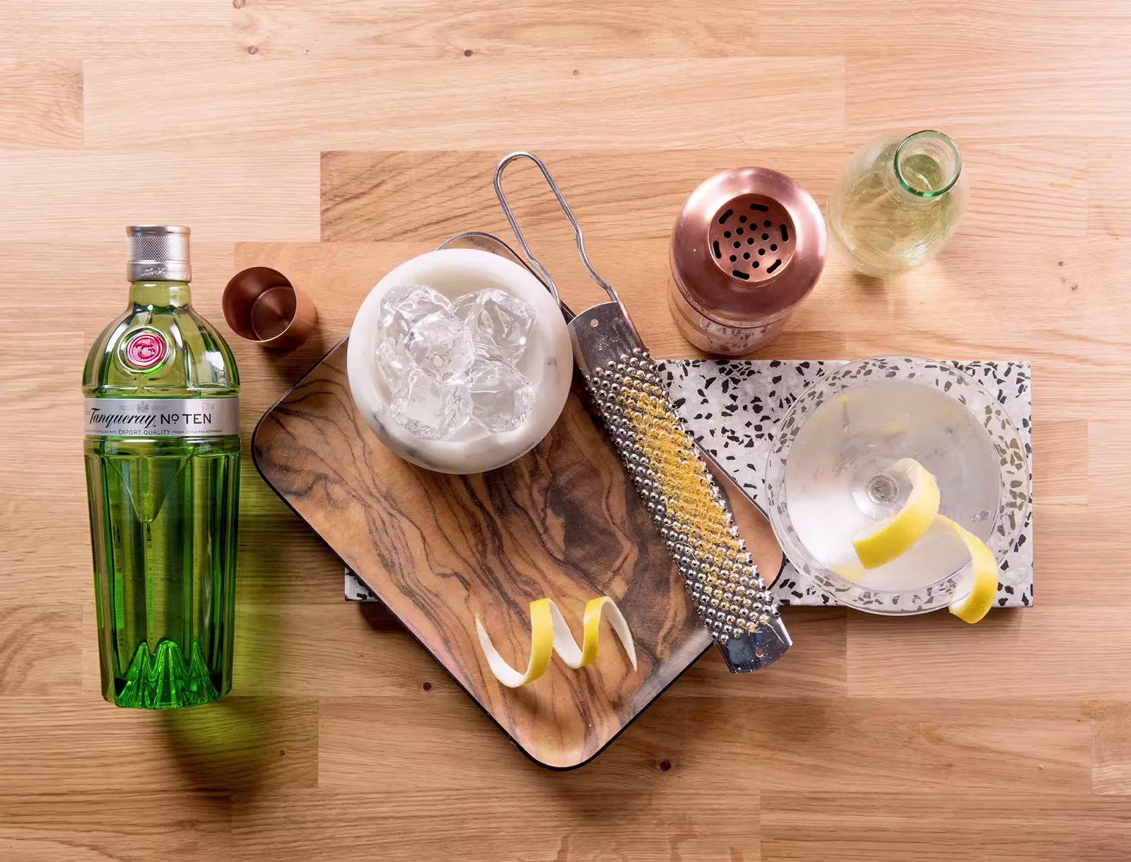 A bottle of Tanqueray gin, a cocktail shaker, a glass with ice, a martini glass with a lemon twist, a grater, and a bottle of tonic water arranged on a wooden surface.