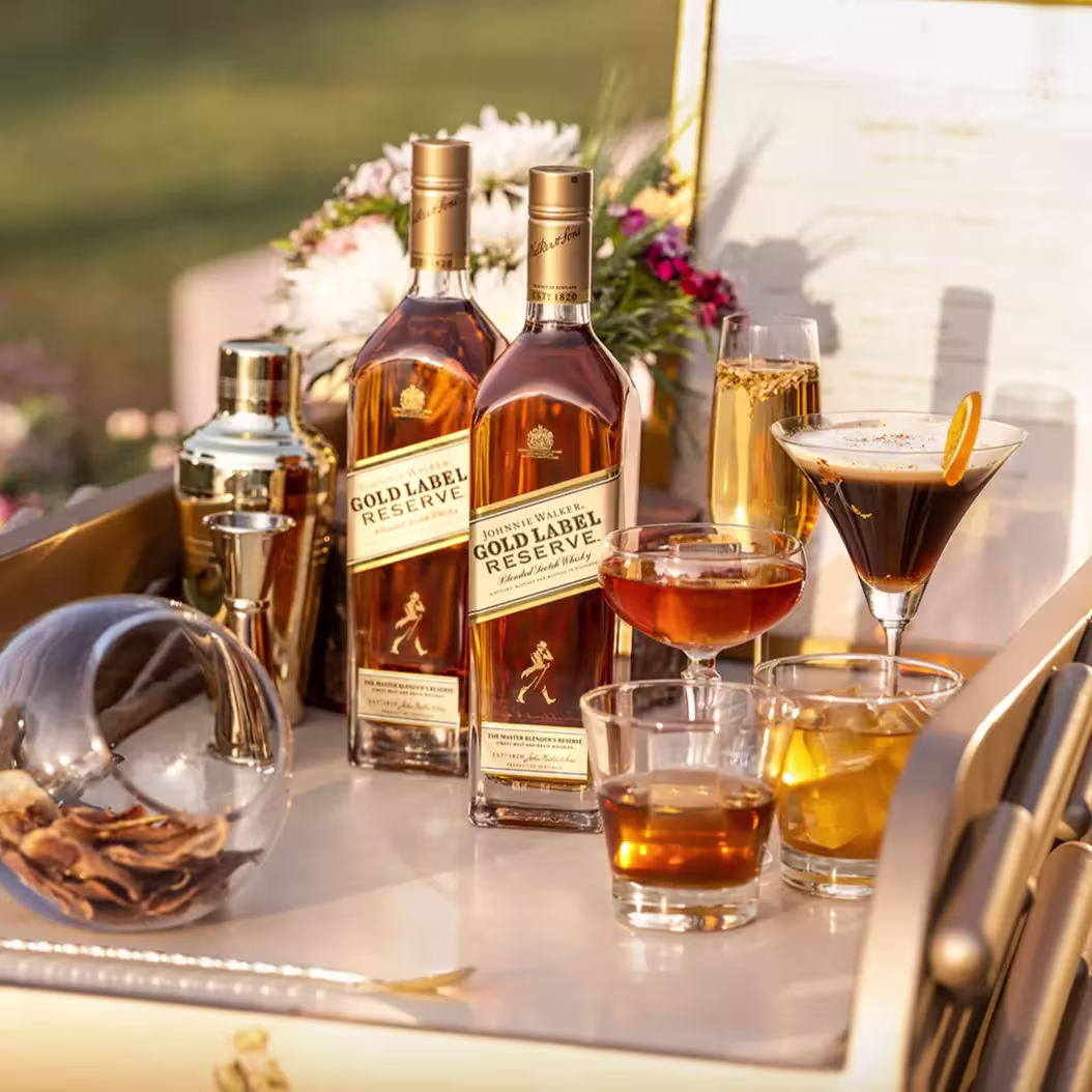 Two bottles of Johnnie Walker Gold Label Reserve whisky on a bar cart with cocktails, a shaker, dried citrus, and flowers in the background.