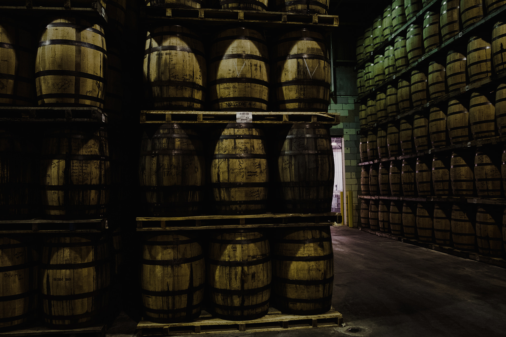 Rows of wooden barrels stacked on pallets in a dimly lit warehouse, with a doorway visible in the background.