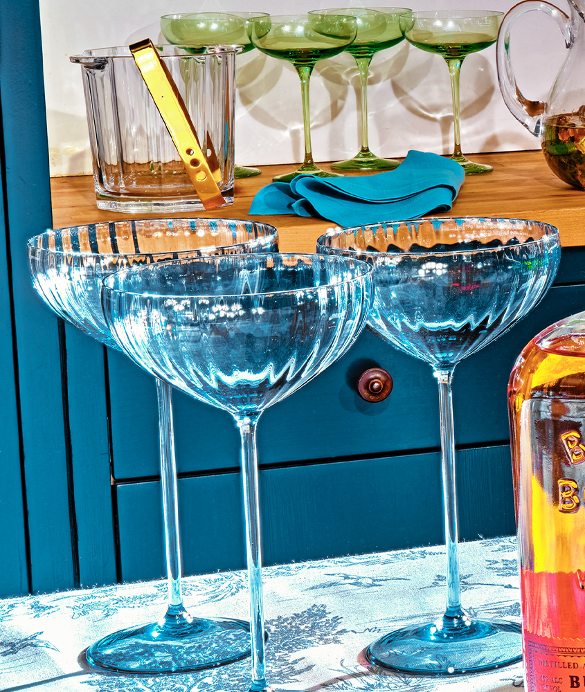 Three clear glass cocktail coupes are arranged on a blue surface in front of a cabinet, with more glasses, a glass ice bucket, and a folded napkin in the background.