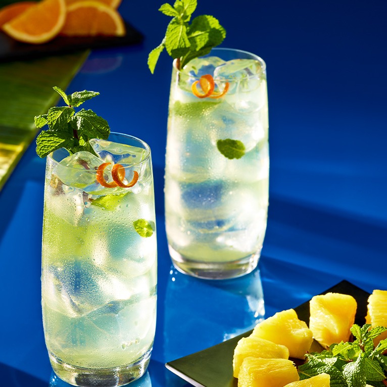 Two mojito cocktails on a blue table next to a plate of cut pineapple.