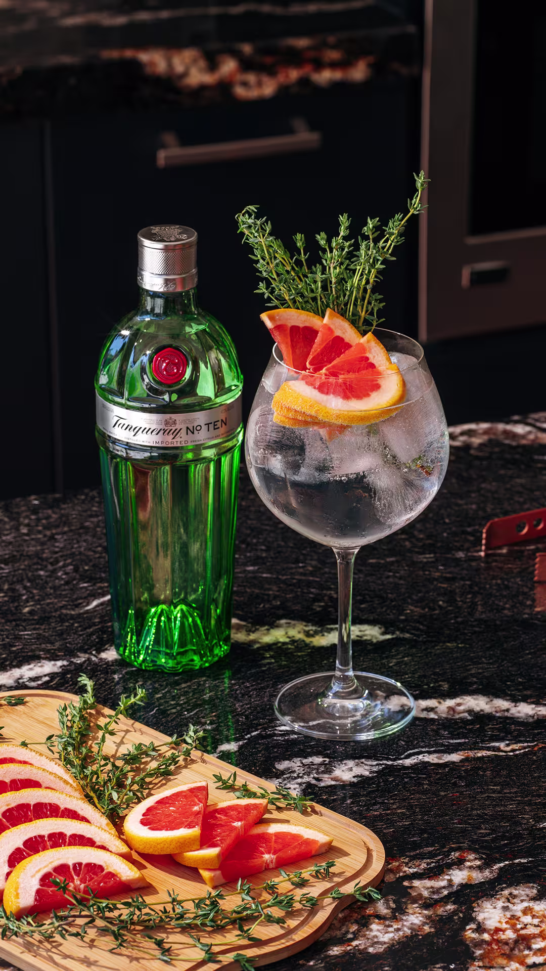 Botella de ginebra Tanqueray No. TEN junto a una copa de gin tonic adornada con rodajas de pomelo y ramitas de tomillo sobre una tabla de madera, con fondo oscuro de cocina.