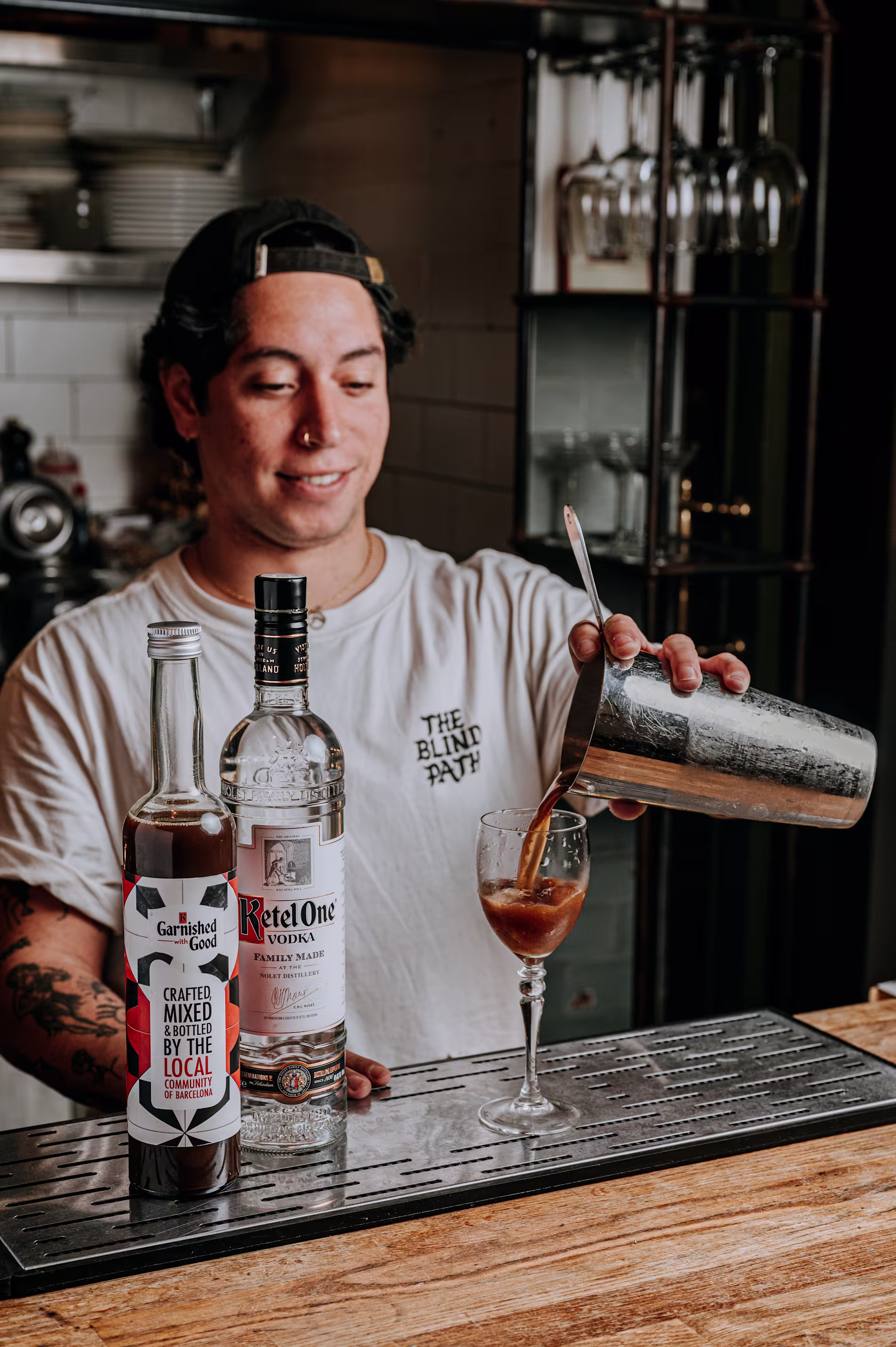 Bartender sirviendo un cóctel desde una coctelera sobre la barra. Junto a él, botellas de Ketel One y Garnished with Good, en un ambiente de cocina con estanterías y copas al fondo.