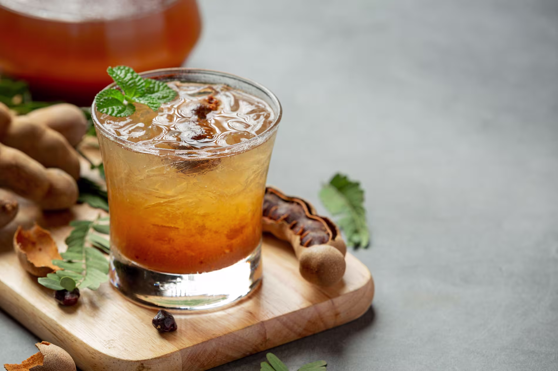 A glass of iced tamarind juice garnished with mint, surrounded by tamarind pods and leaves on a wooden board.