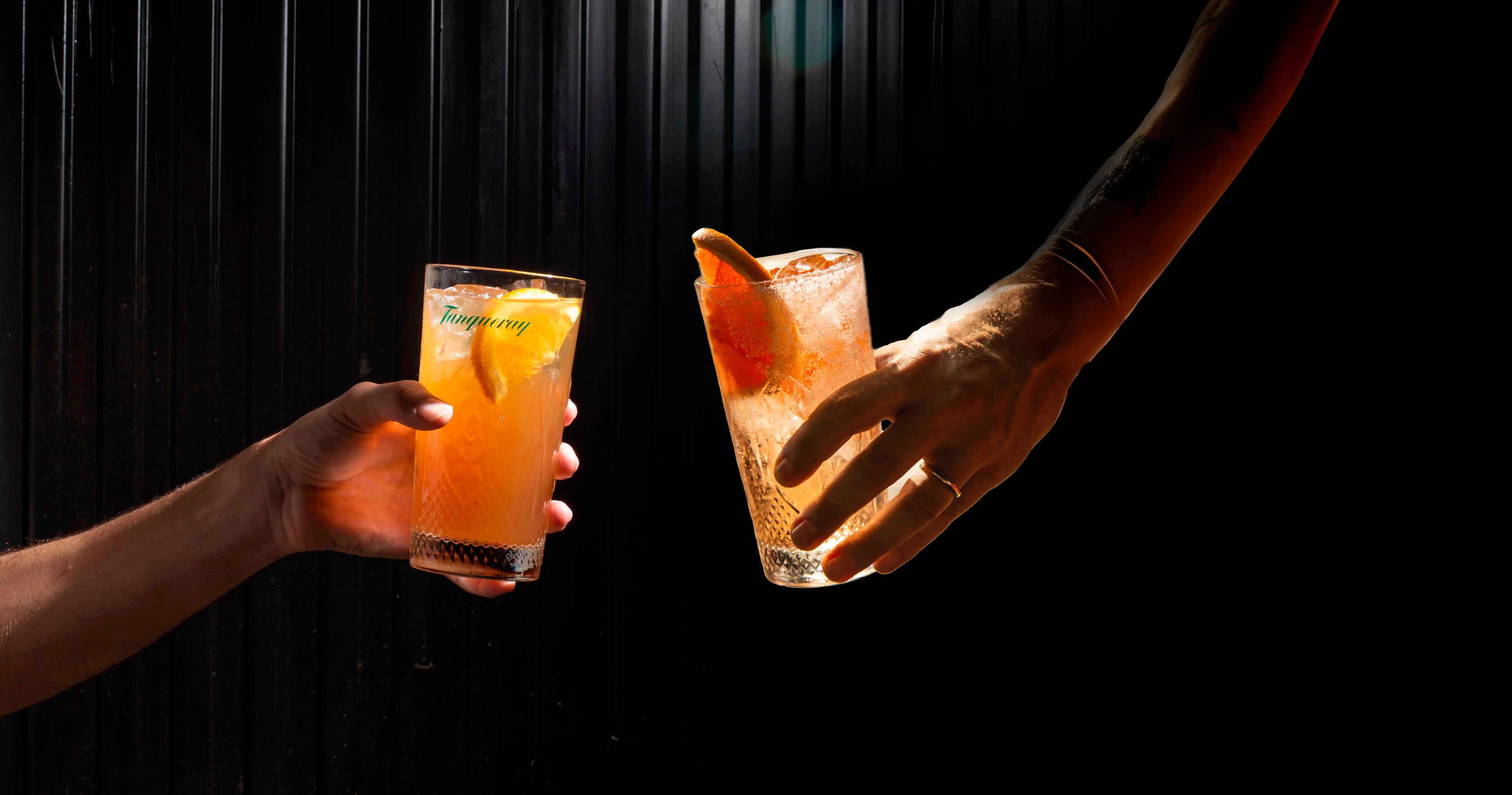 La imagen muestra dos manos, una a cada lado del marco, sosteniendo vasos con un líquido y una rodaja de fruta.